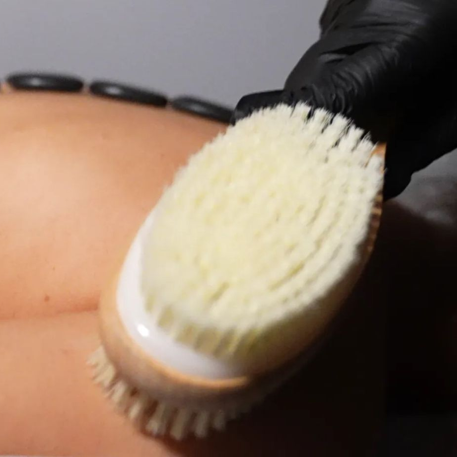 Person's back being dry brushed with a soft brush, with hot stones in place. Hand in black glove.