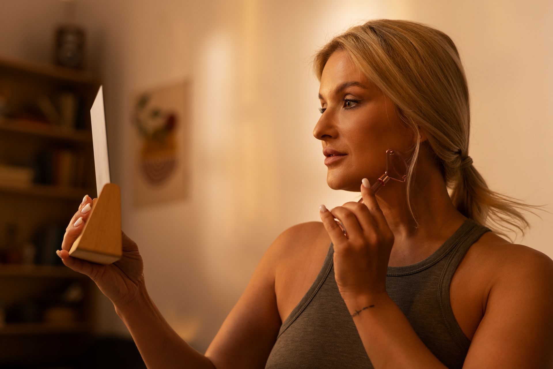 Blonde woman looks at a small mirror while holding a facial tool in her hand; indoors, soft lighting.