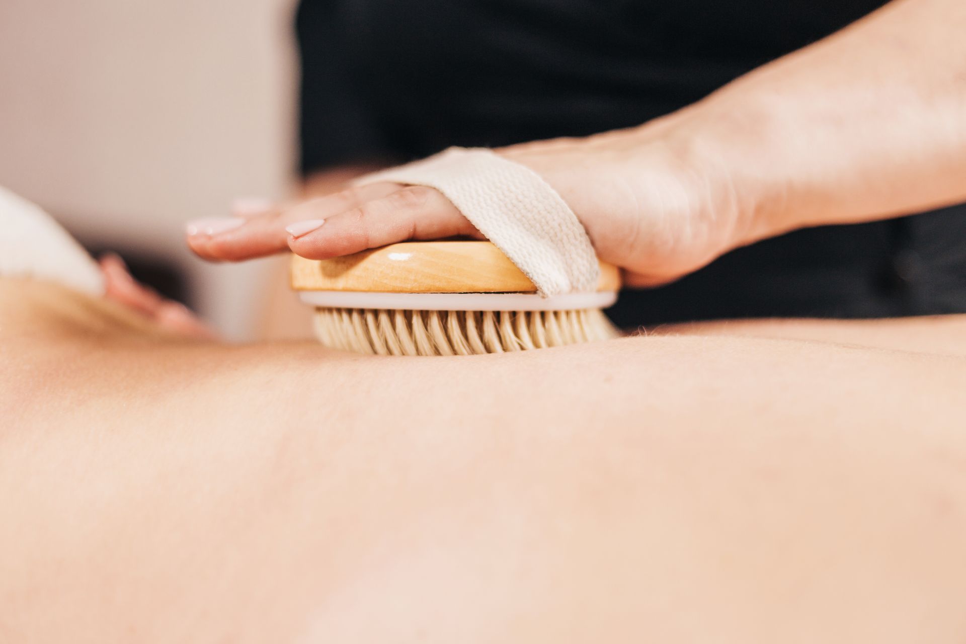 Person dry brushing another's back with a round wooden brush, tan bristles, in a treatment setting.