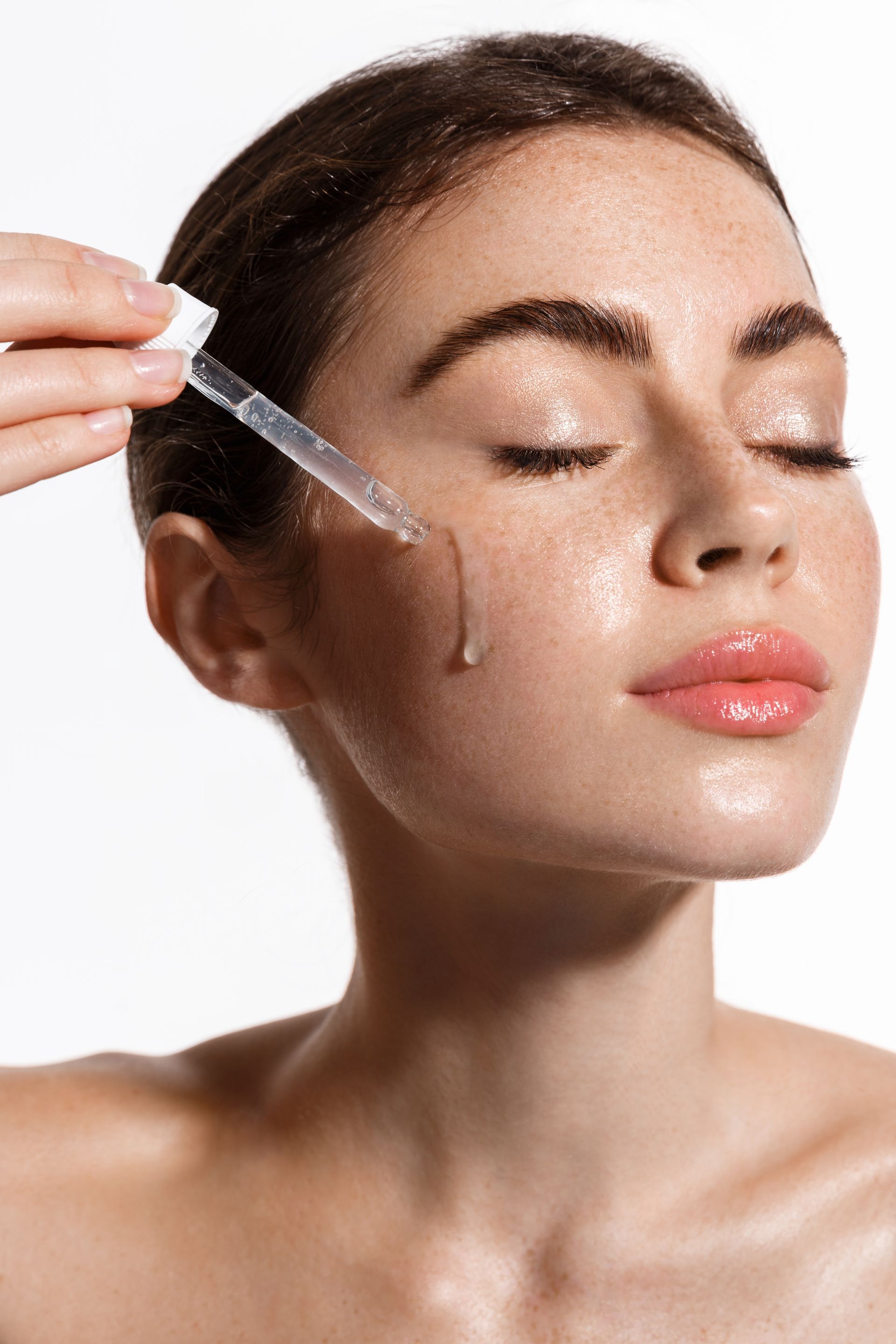 Woman applying serum with dropper; eyes closed, glowing skin, against white background.