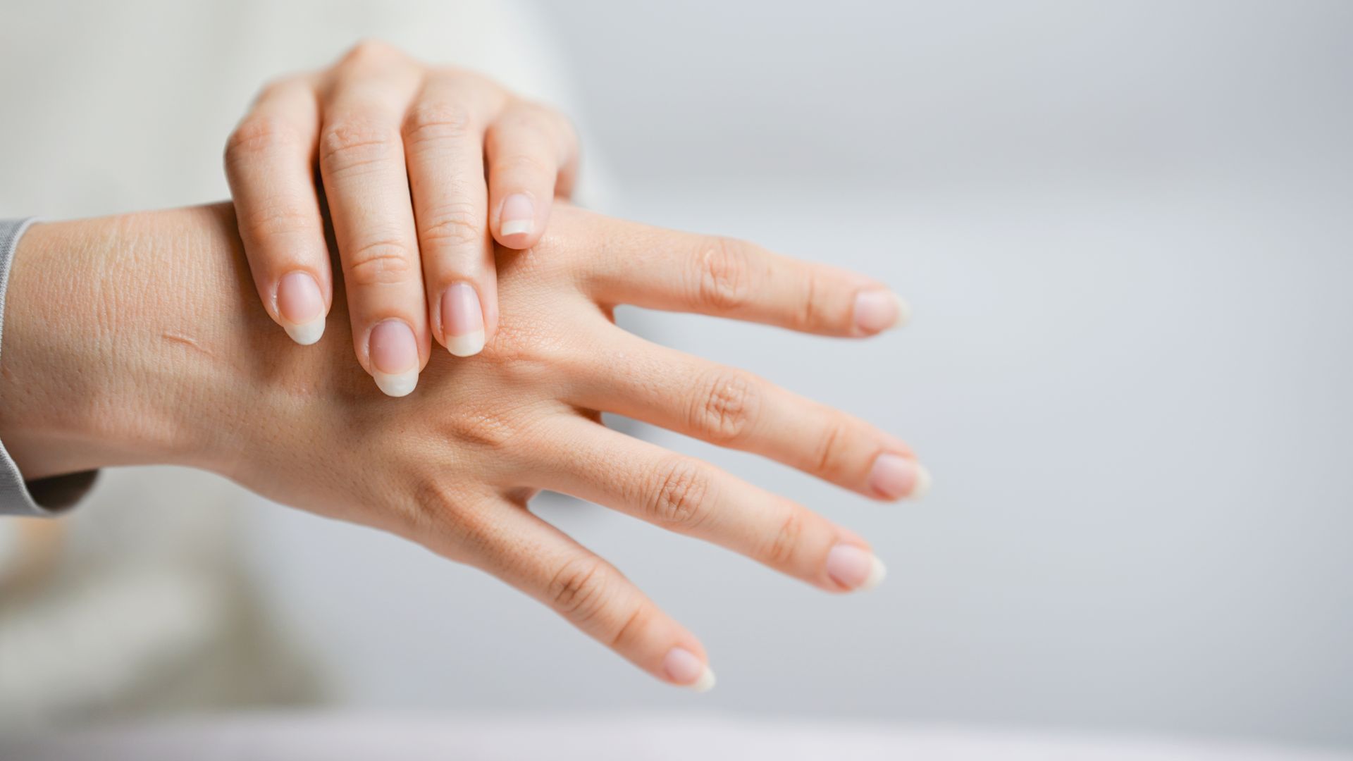 Person’s hands, one resting on the other. Fingers extended, showing nails. Neutral background.