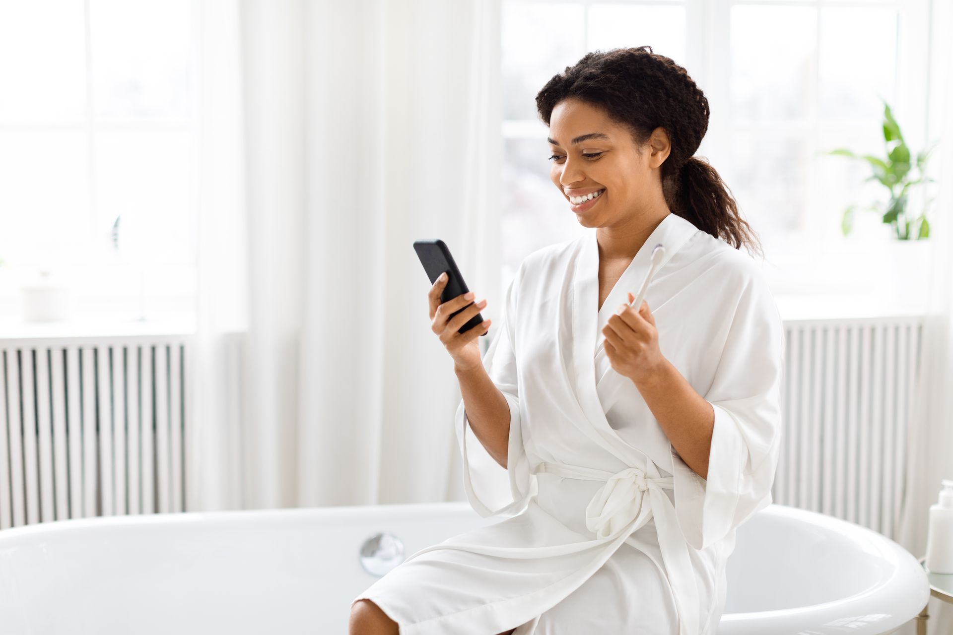 Woman in a white robe smiles, looking at her phone in a bright bathroom, sitting on the edge of a bathtub.