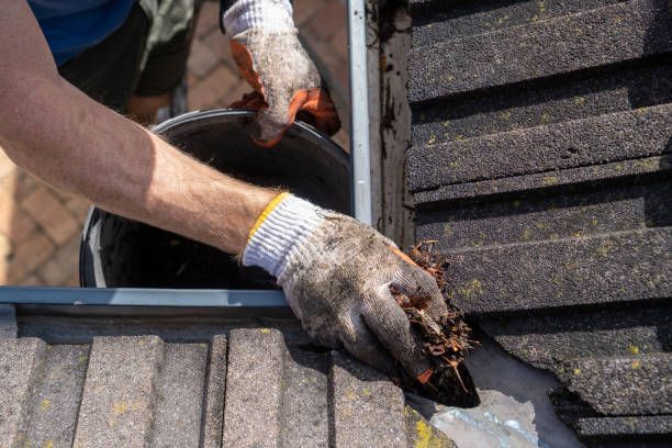 A close-up view of gloved hands removing debris from a roof gutter.