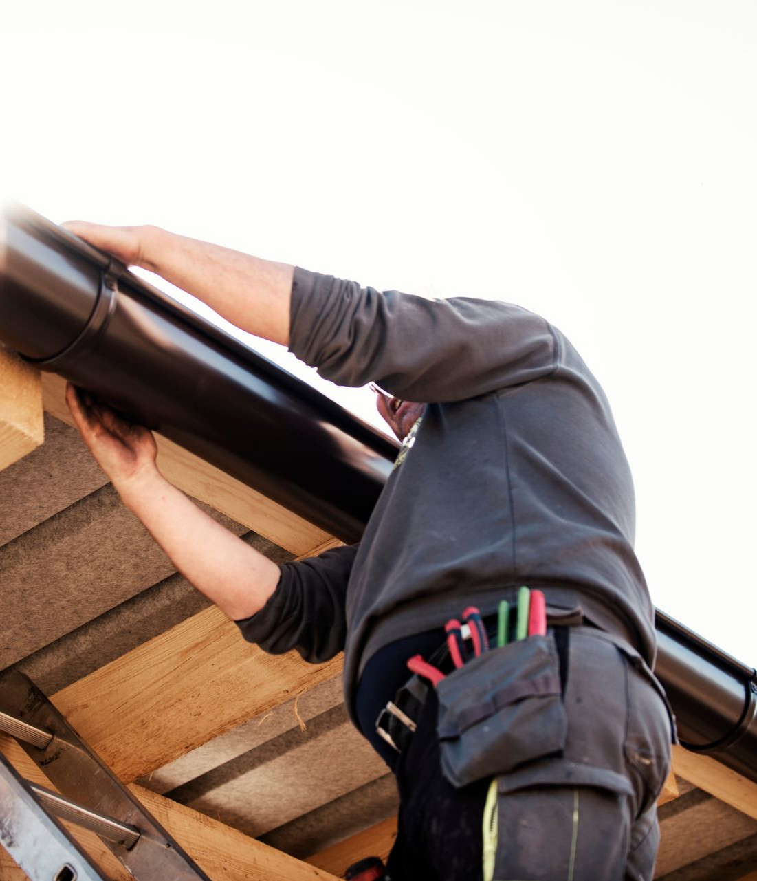 A male worker installs a black gutter on the roof of a wooden house, under the sun.