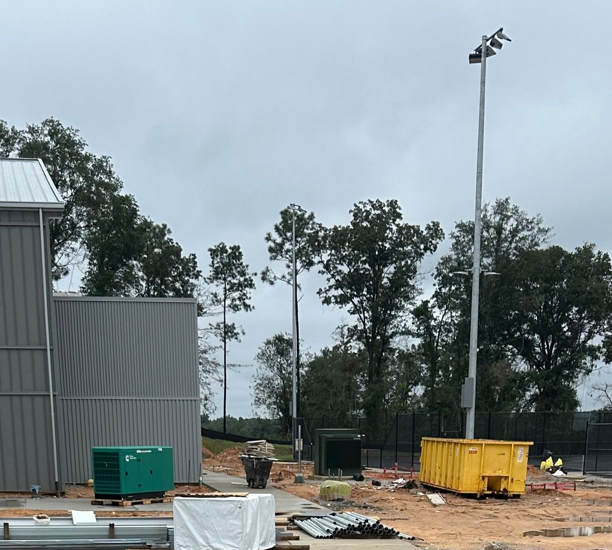 Construction site with metal buildings, tall light pole, and a yellow dumpster against a cloudy sky.