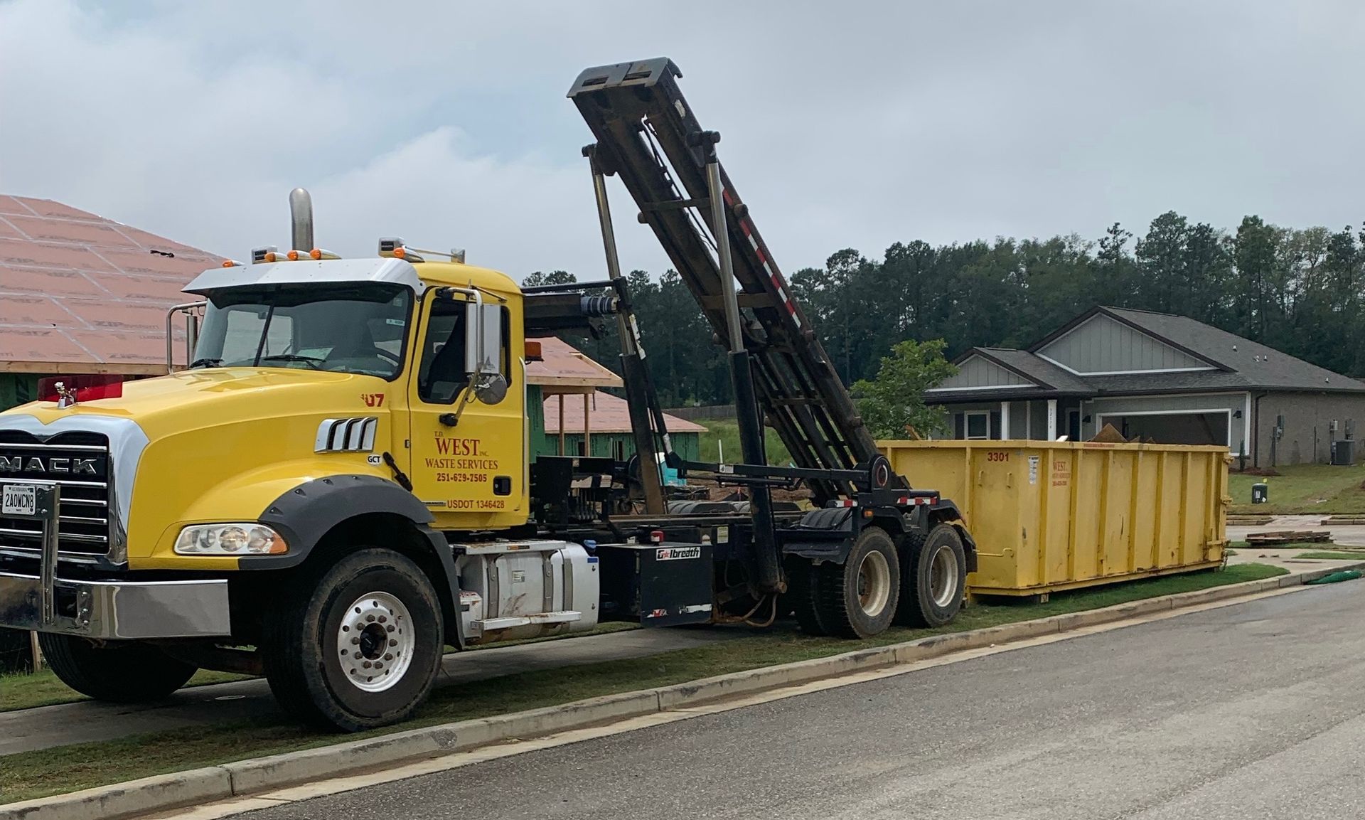 Yellow Mack truck with raised arm is next to a yellow dumpster, on a residential street.