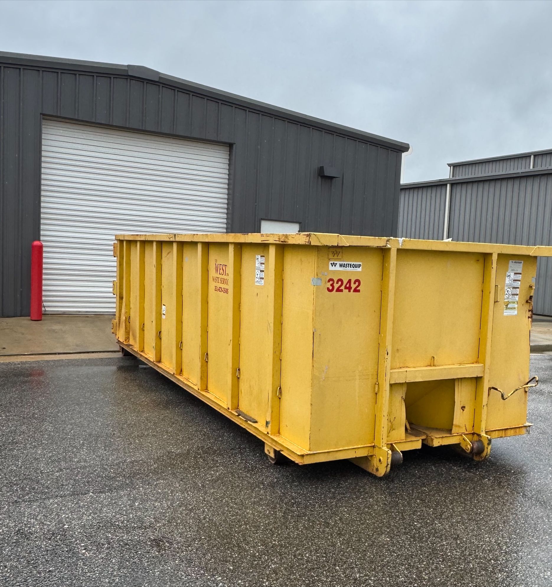 Yellow dumpster in front of a gray building with a roll-up door.