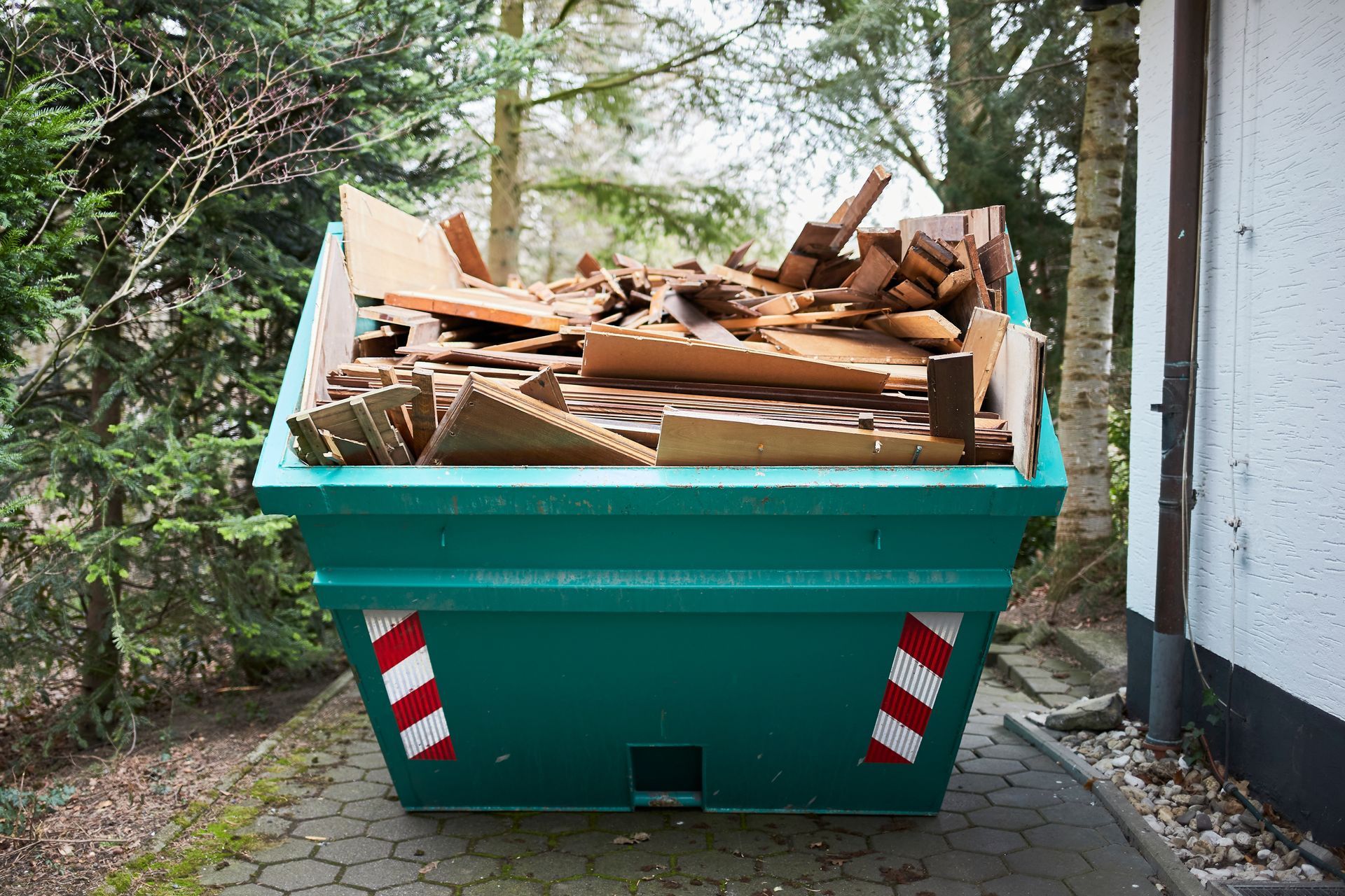 Green dumpster filled with wooden debris, sitting on a paved driveway near a building and trees.