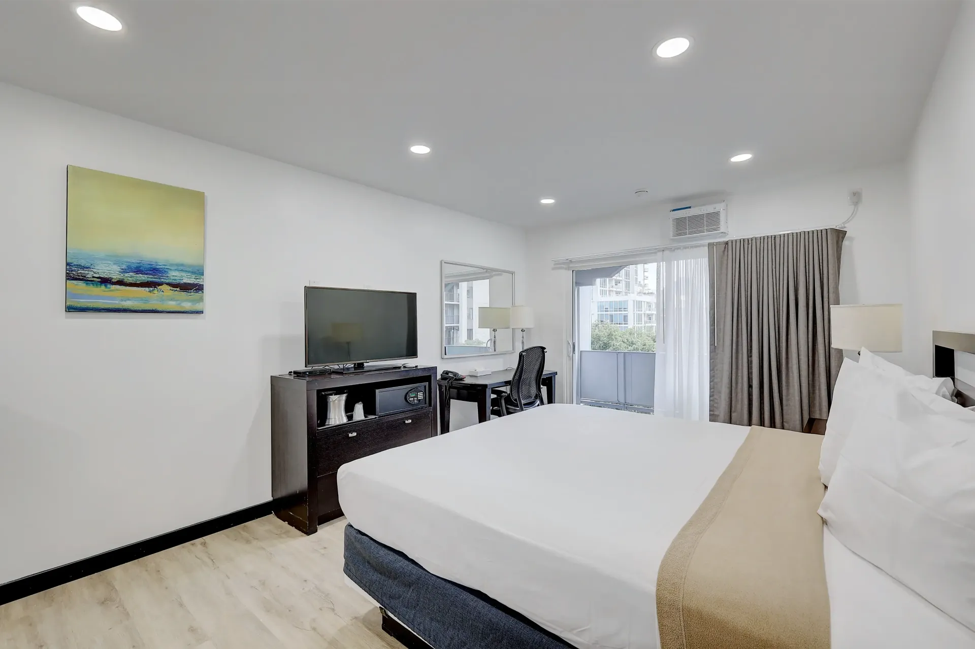Bedroom with a bed, TV, desk, and balcony access. White walls, light wood floor, and a colorful painting.
