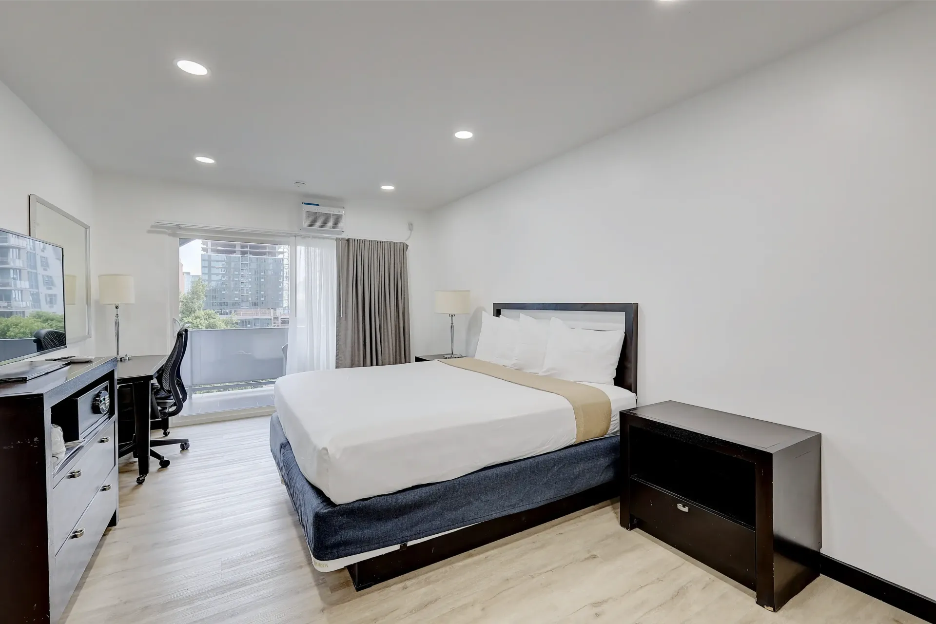 Bedroom with a bed, desk, and balcony. White walls and floors, dark furniture.