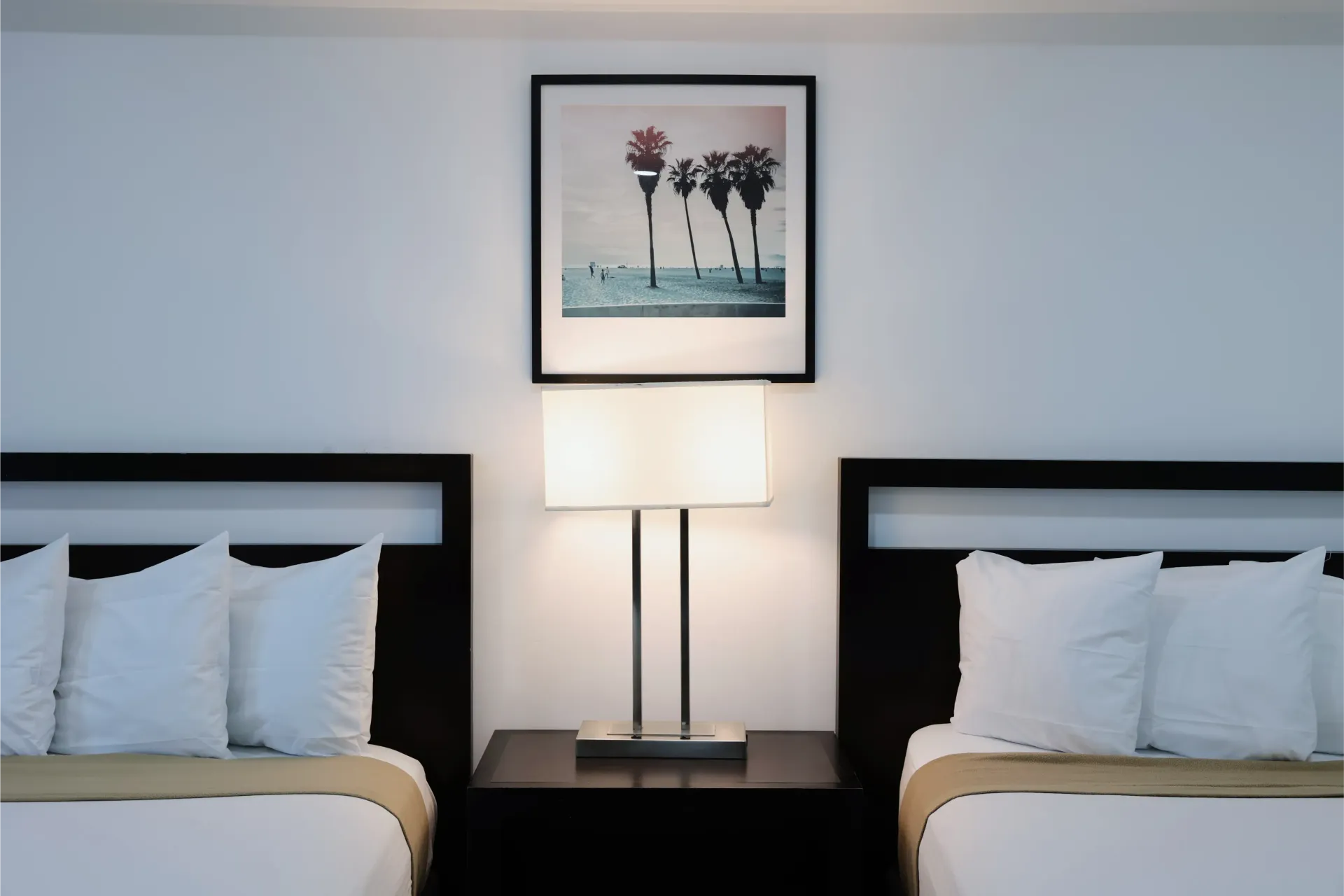 Two beds with white linens, nightstand with lamp, and framed art on white wall.