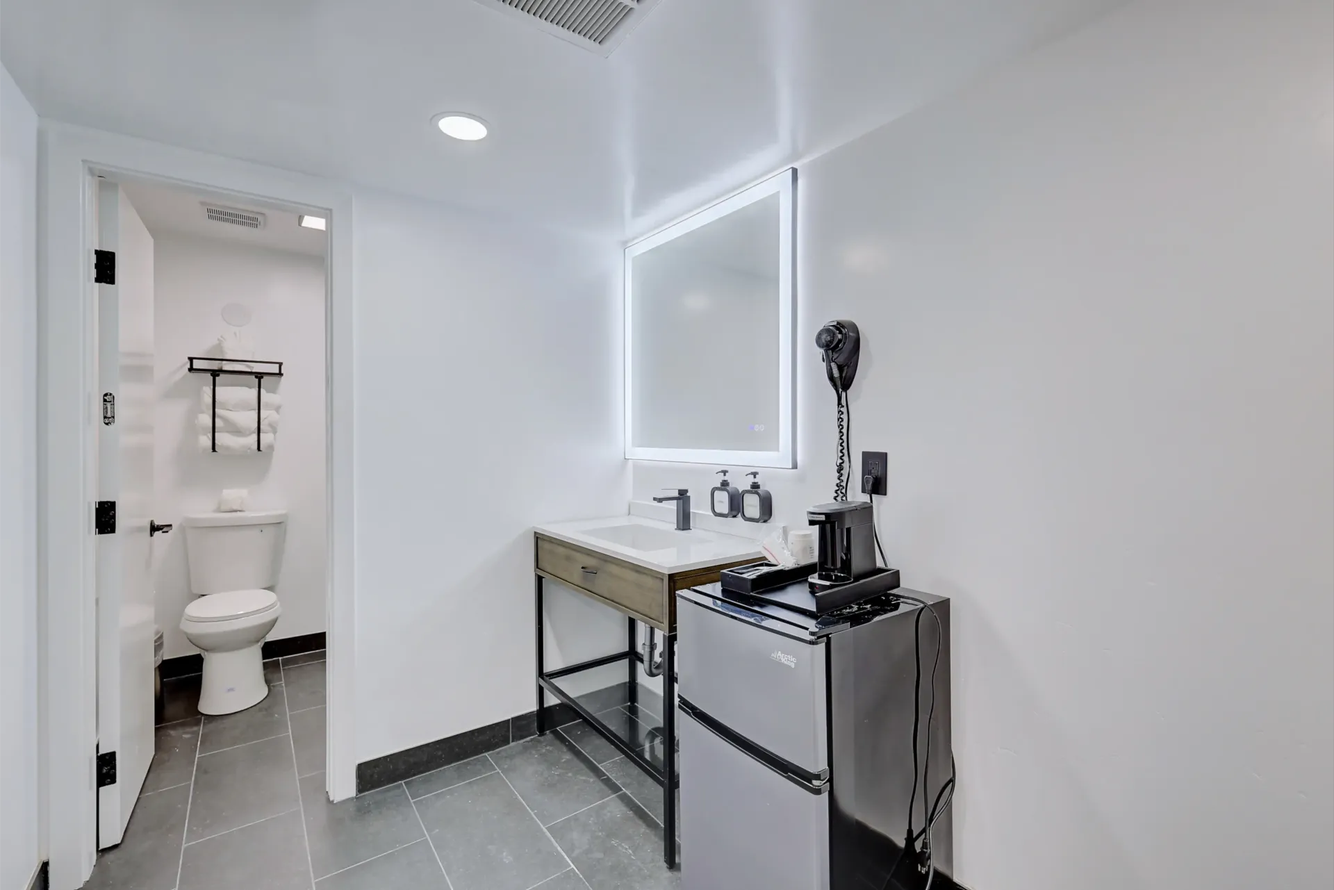 Bathroom with toilet, vanity, and mirror. White walls, grey floor, and a silver machine.