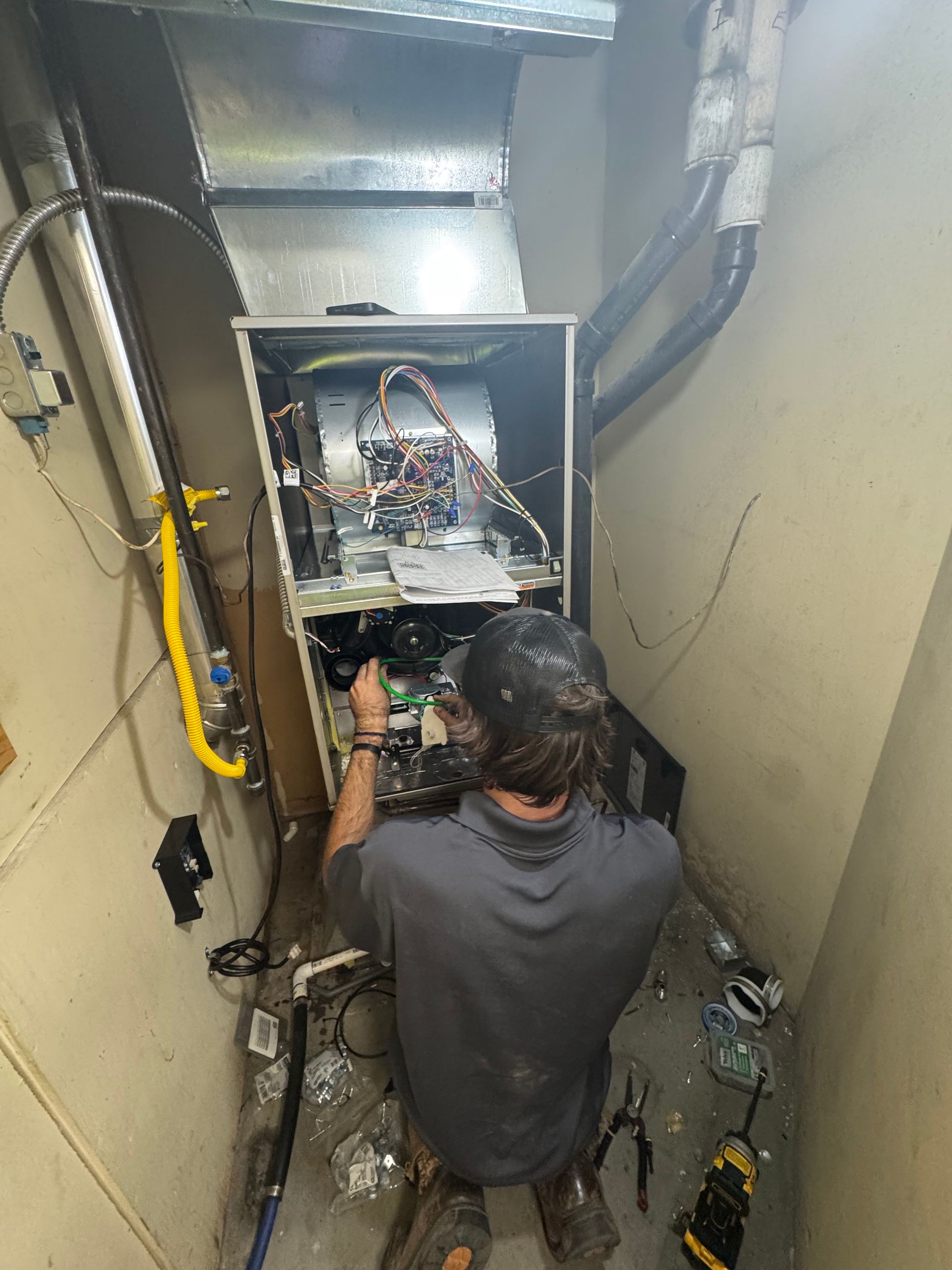 A person repairs an HVAC unit in a tight space. They are kneeling, working on the open unit, with tools and parts scattered around.