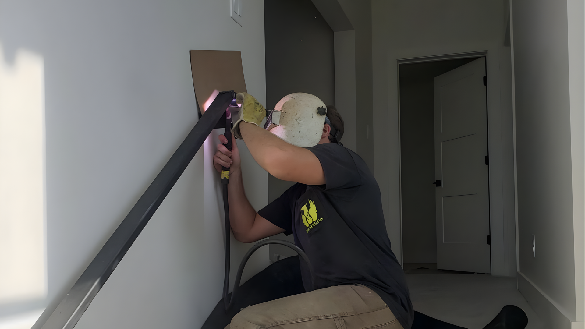 Person kneeling on a stairwell, using a tool to repair a wall corner beside a handrail.