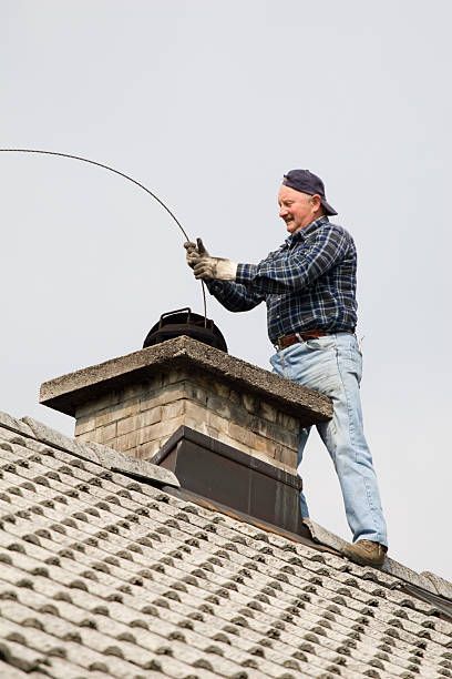 A man is standing on top of a roof cleaning a chimney.
