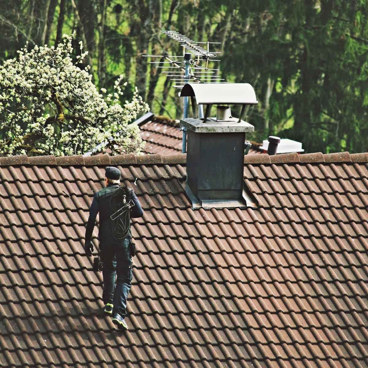 A man is standing on a tiled roof with a chimney in the background.