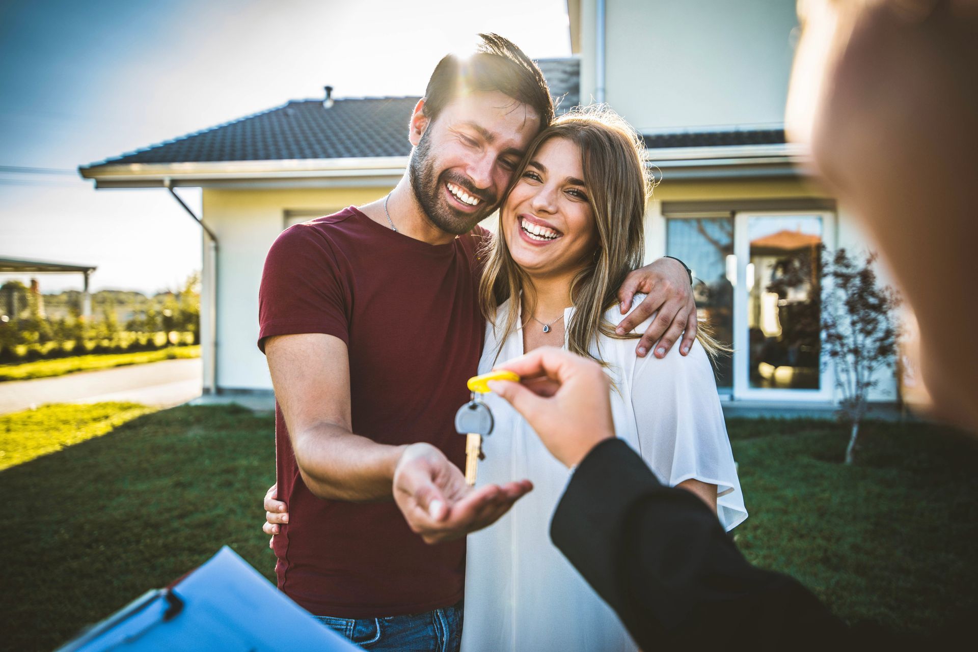 Couple smiling outside a house as a hand offers them keys to a new home
