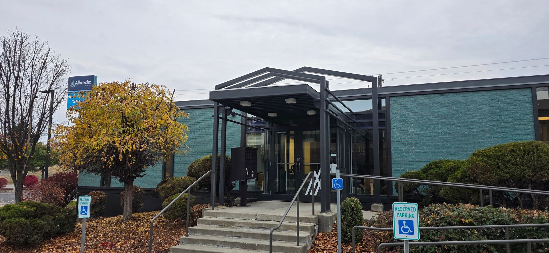 Accessible building entrance with glass doors, steps, autumn trees, and a handicap parking sign
