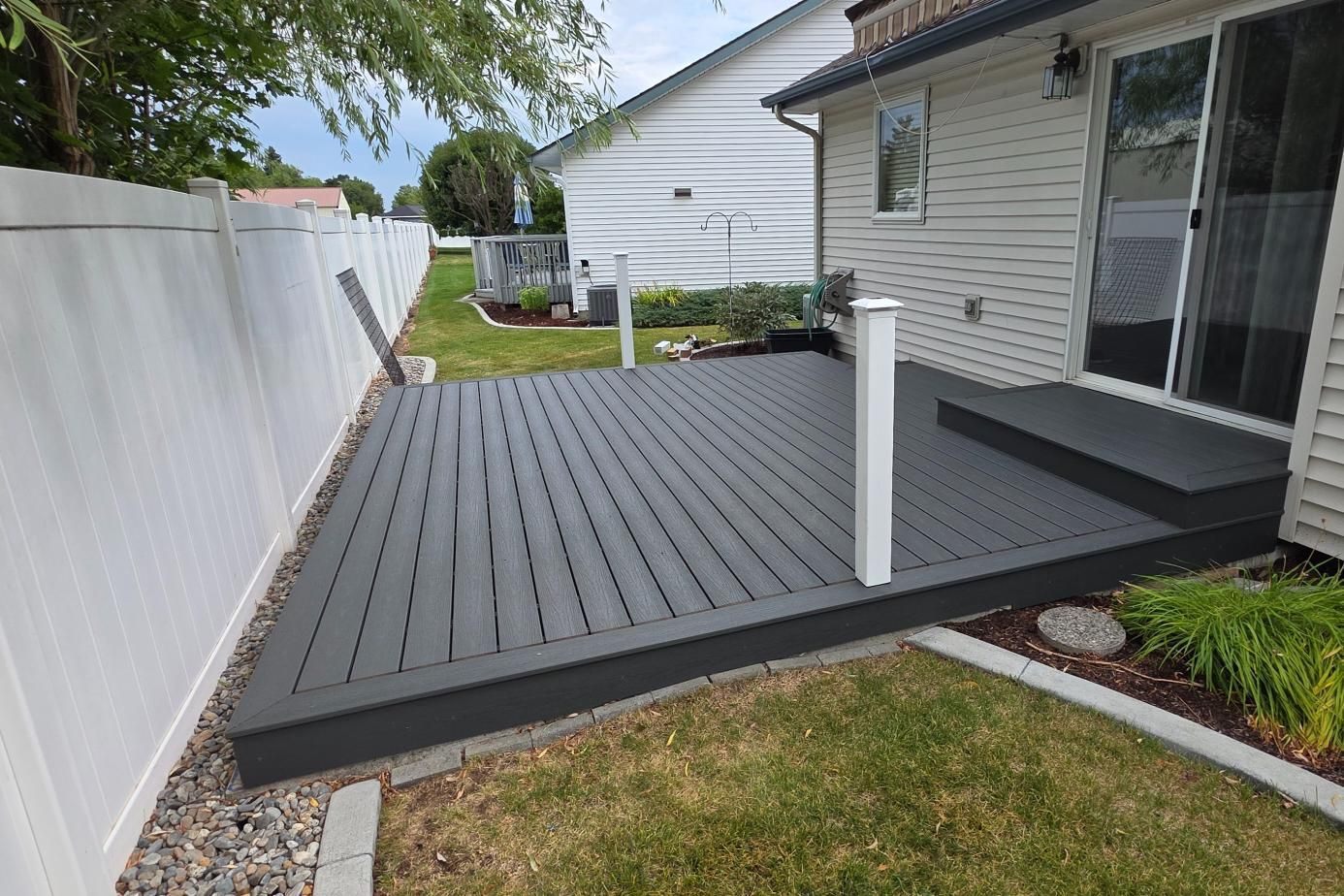Raised gray deck beside a house and white fence, with a small white post and sliding glass door.