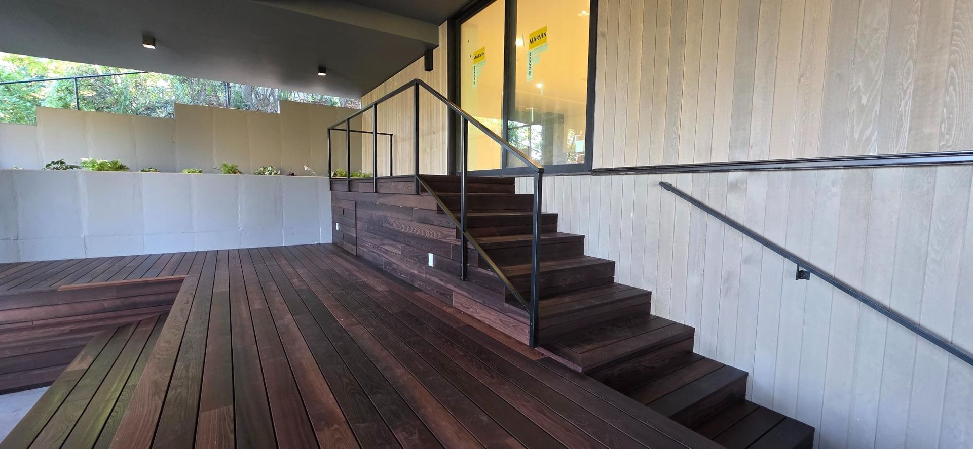 Wooden deck with stairs and black railing leading to a building entrance and white planter wall