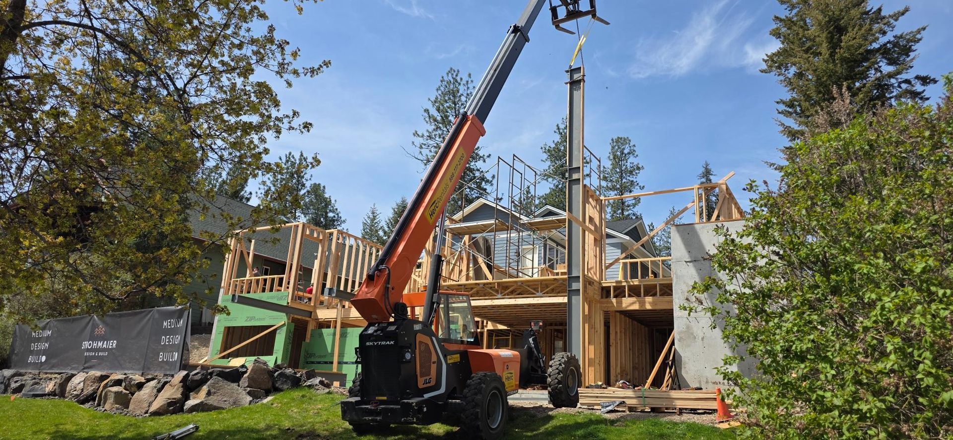 Construction site with crane lifting materials beside a partially built wooden house in a wooded area