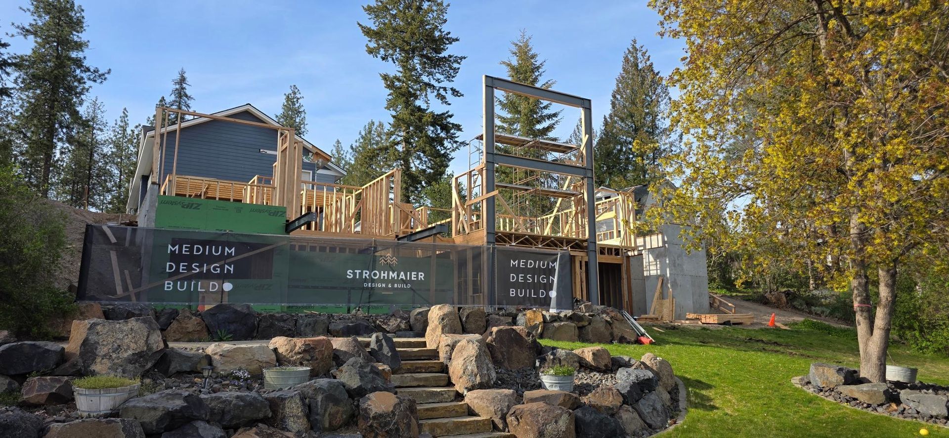 Mountain lodge under construction with wooden framing, green tarp, and a stone garden in the foreground