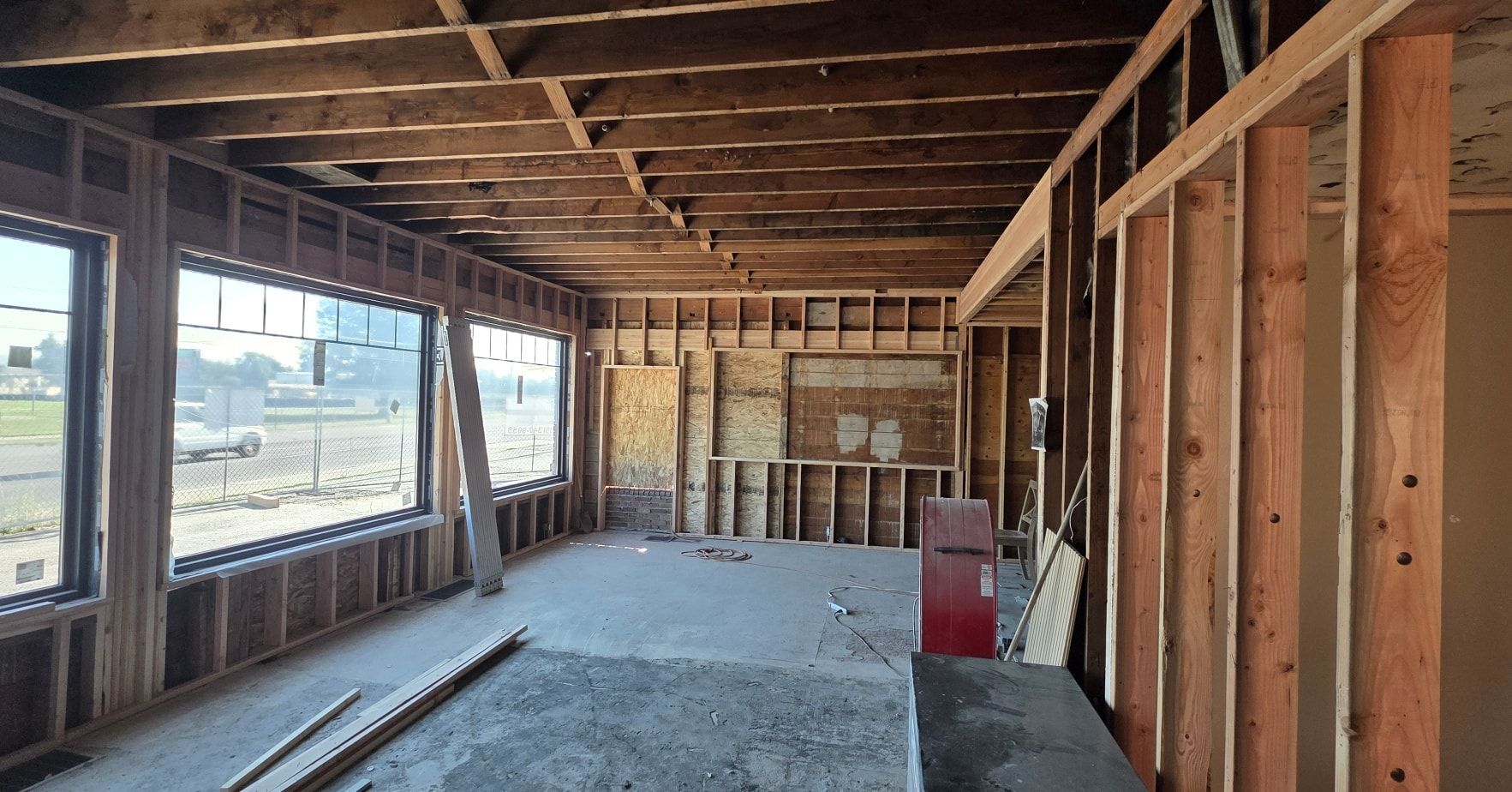 Interior of a house under construction with exposed studs, unfinished floor, and large windows on the left.
