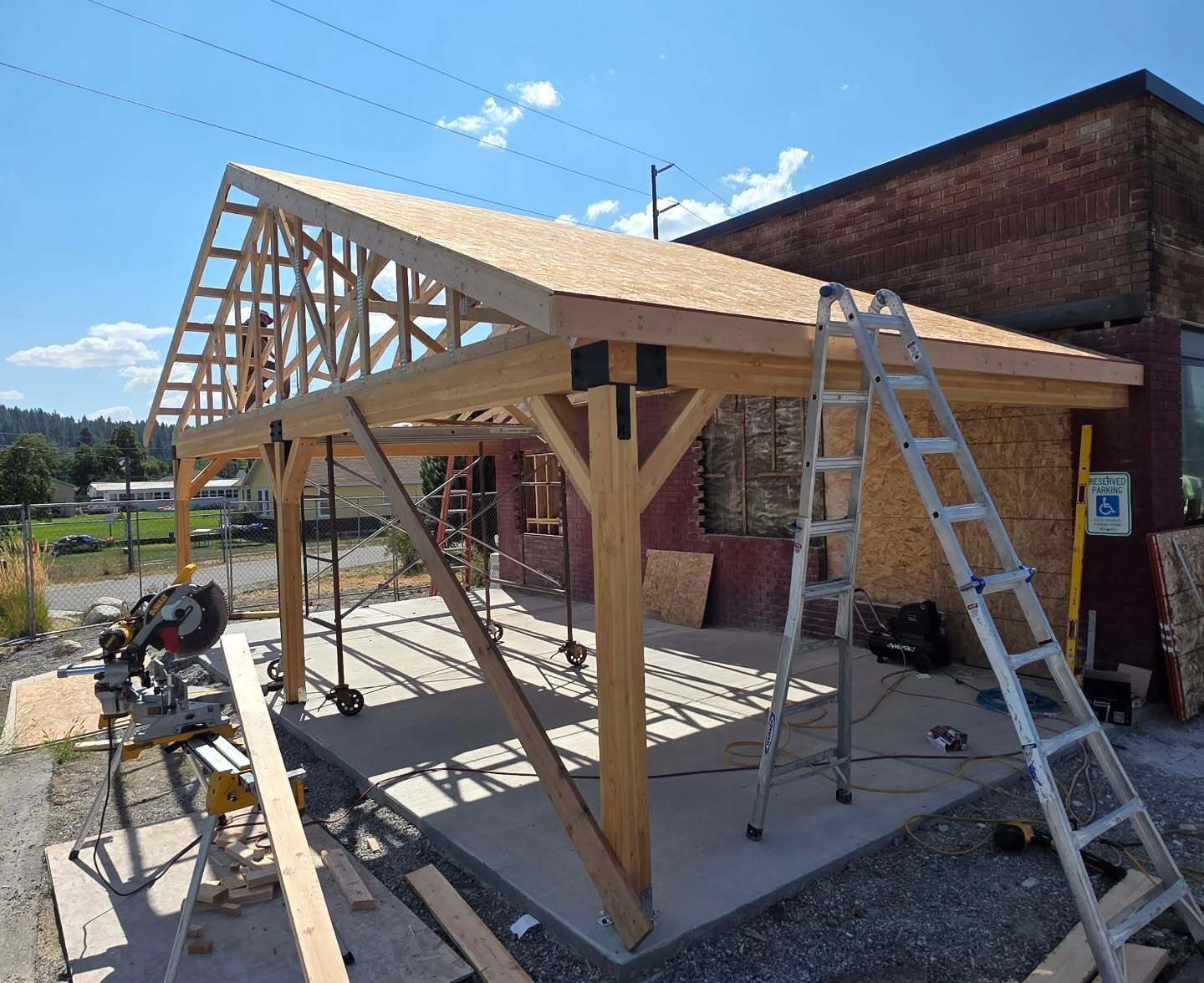 Partially built wooden carport frame beside a brick building, with ladders under a sunny sky