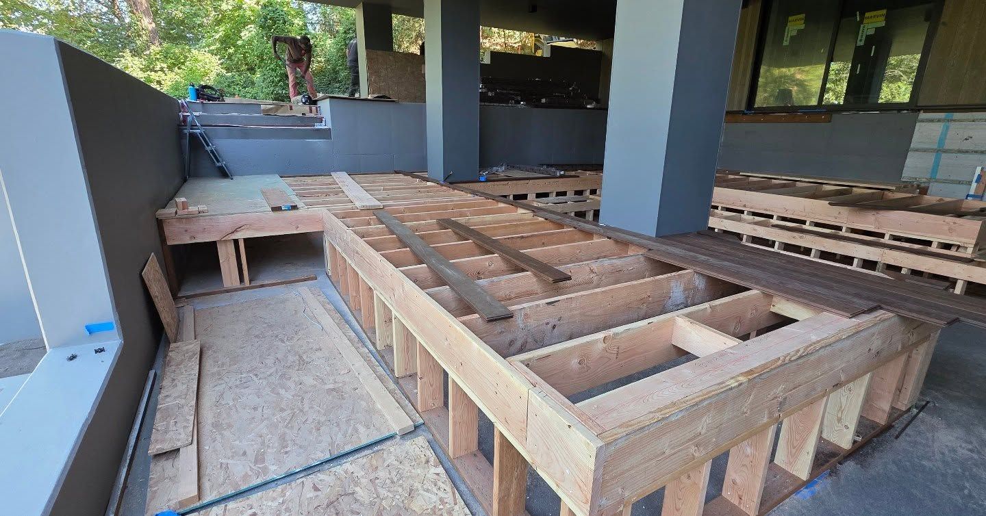 Raised wooden deck frame under construction on a covered patio, with blue support columns and stone flooring.