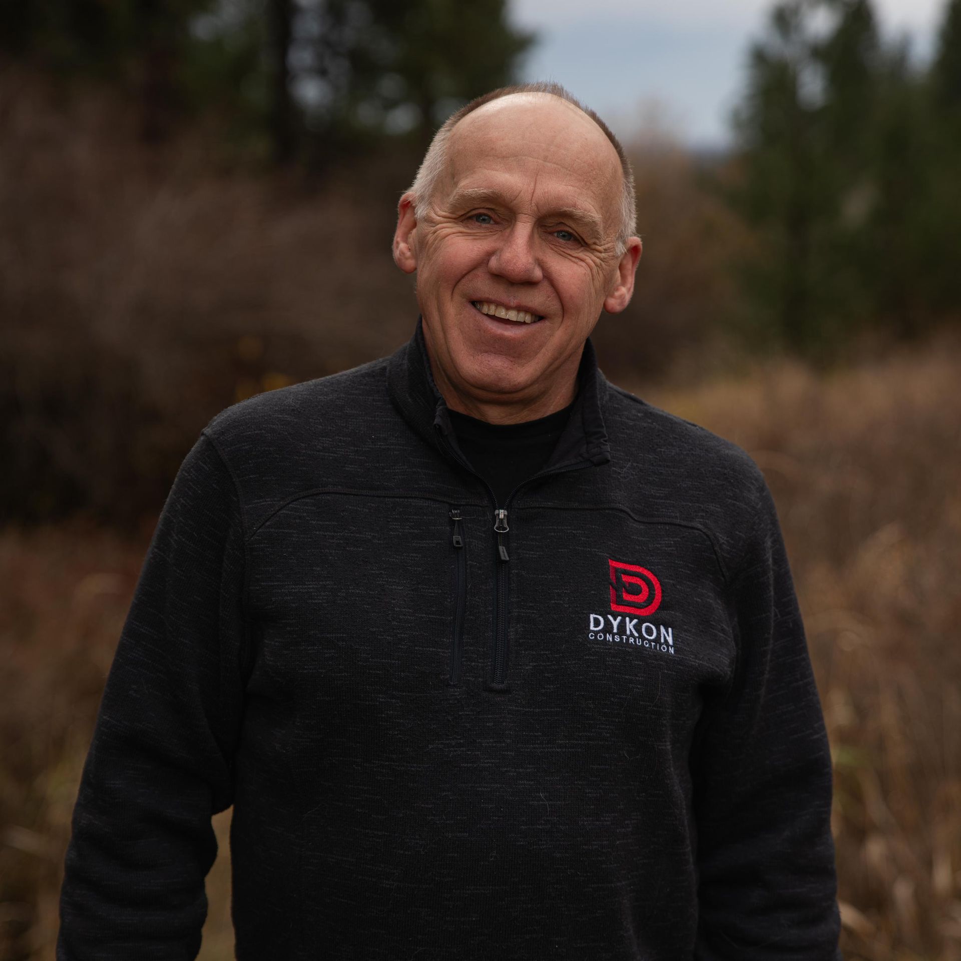 Smiling man in a black fleece jacket standing outdoors with blurred trees behind him