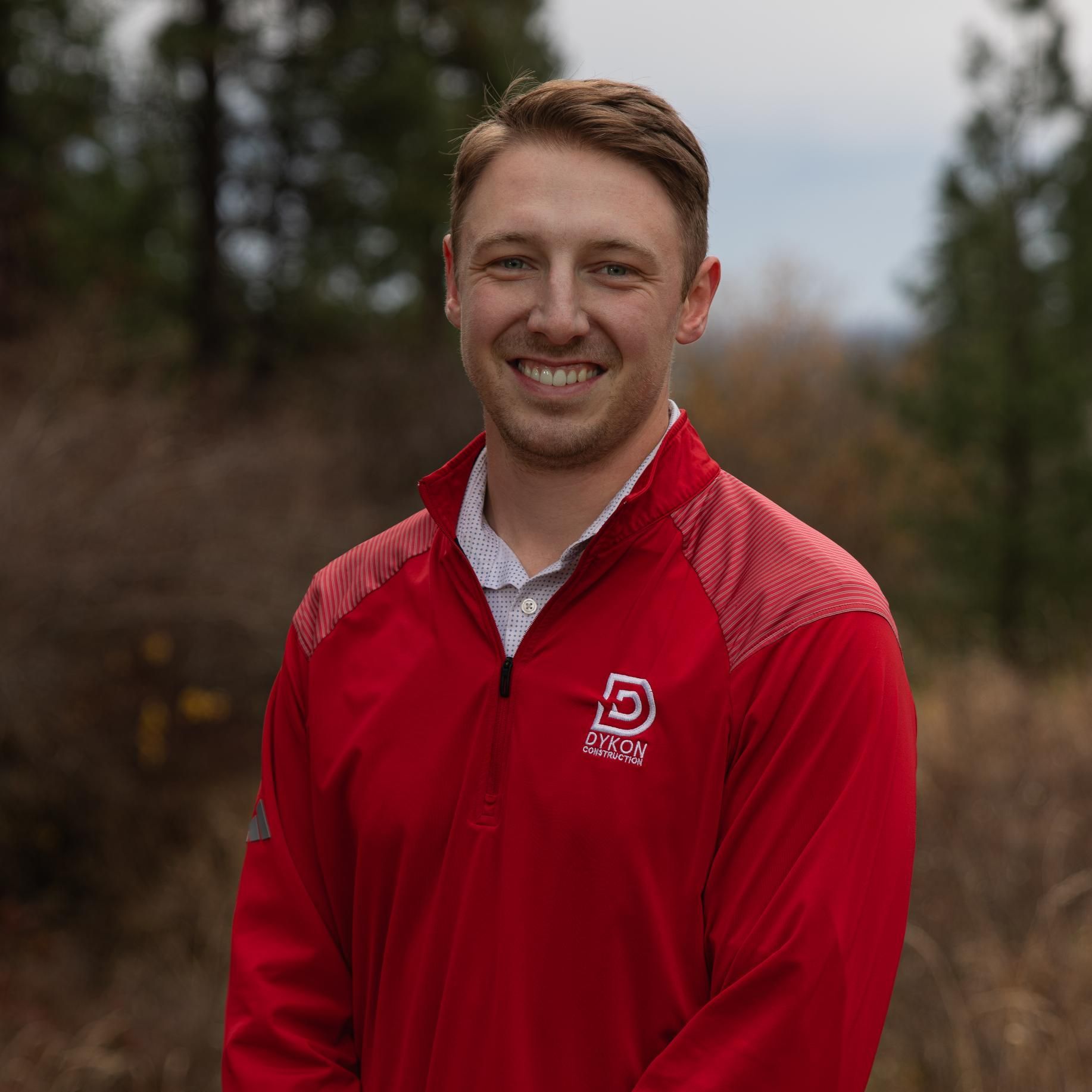 Man in a red zip-up jacket standing outdoors, smiling in front of a blurred wooded background
