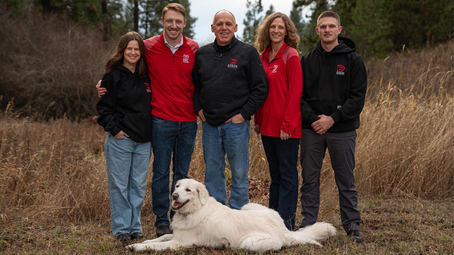 Five people stand outdoors in red and black jackets with a white dog lying in front of them.