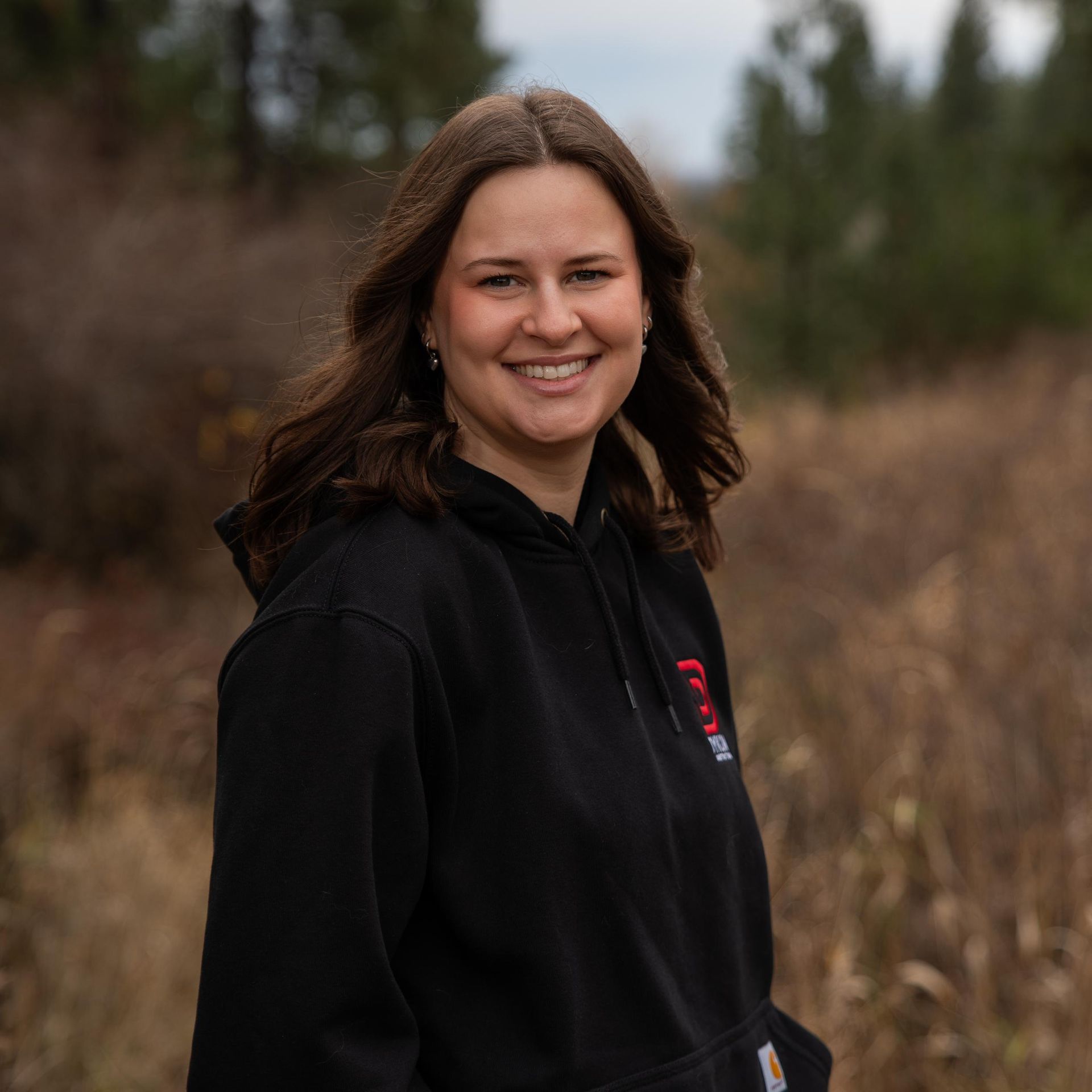 Smiling person in a black hoodie standing outdoors in a grassy field with trees in the background