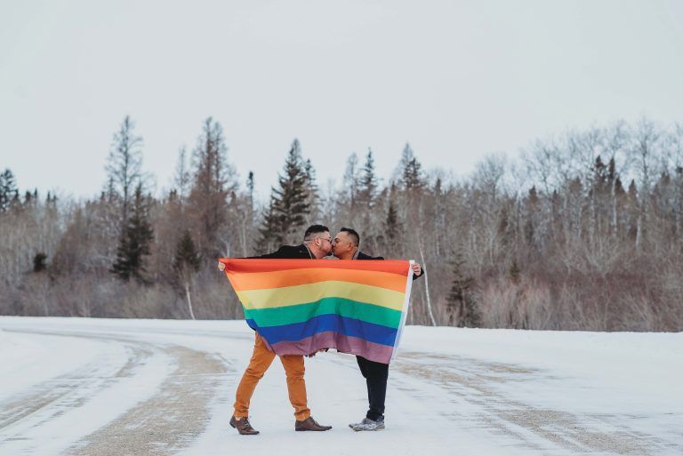 Two men are kissing while holding a rainbow flag in the snow.