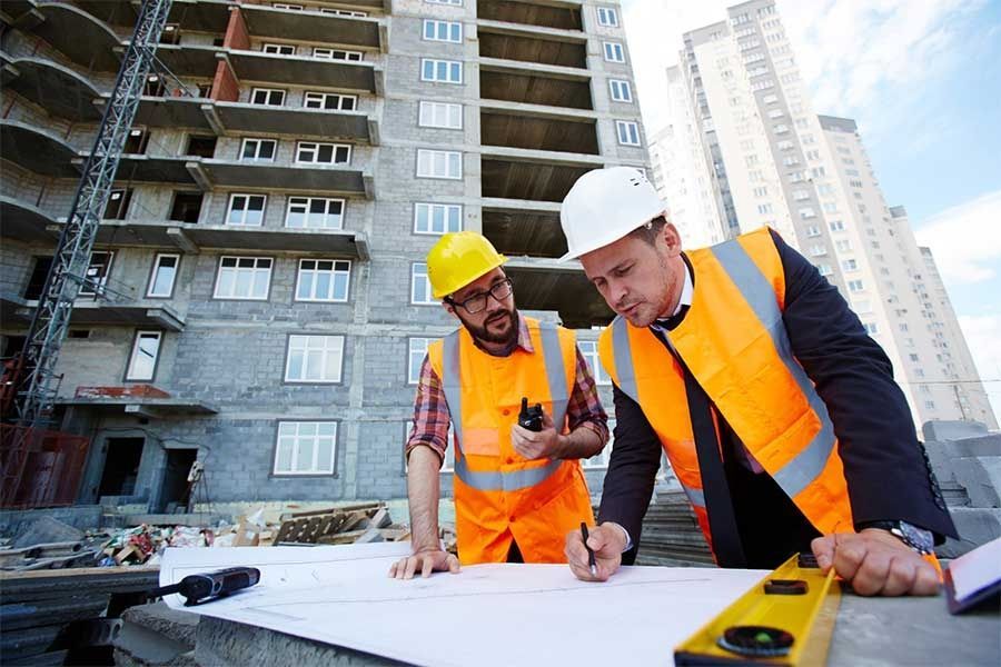 Construction workers in orange vests reviewing blueprints at a building site with high-rise apartments behind them