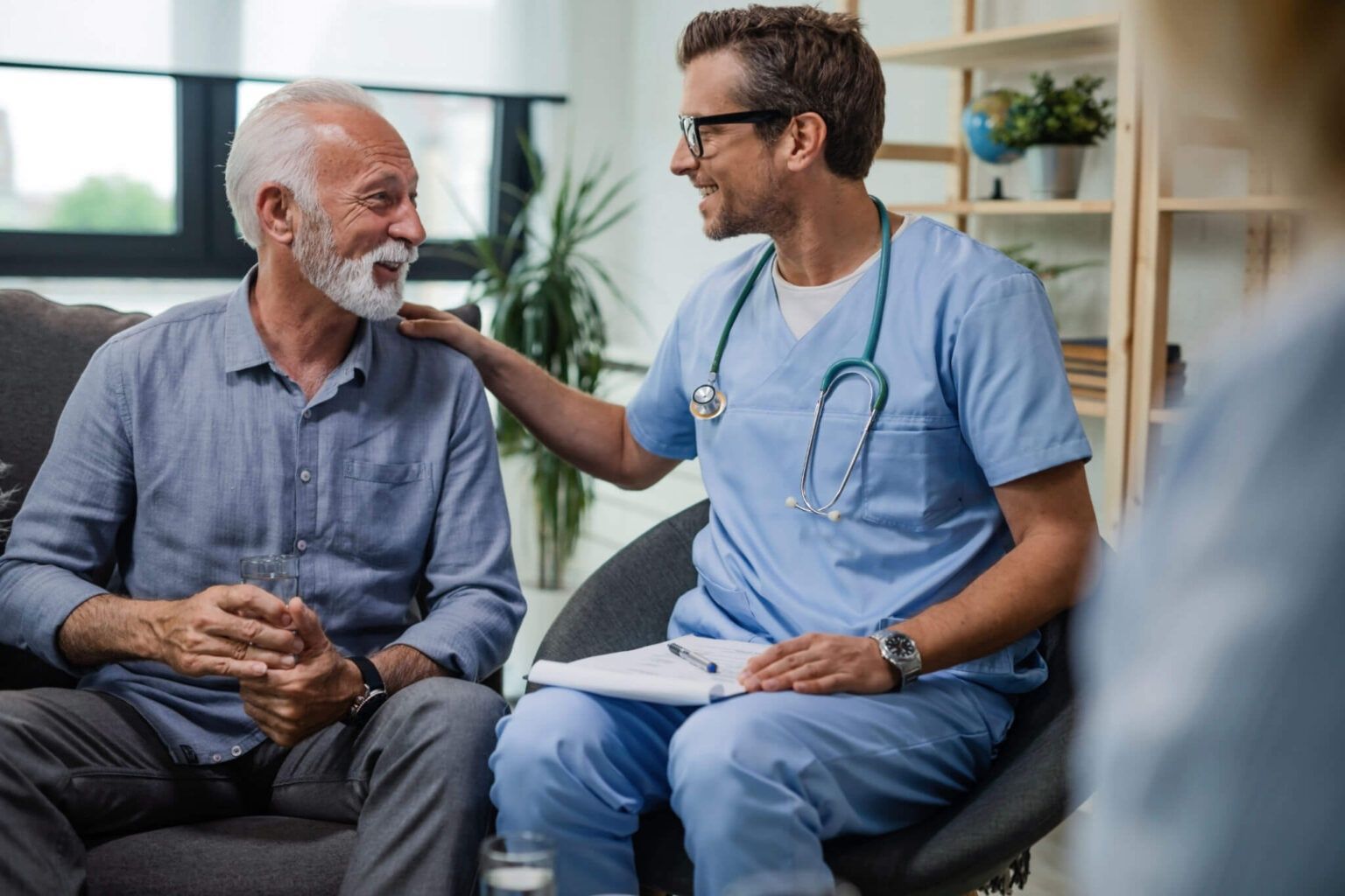 Doctor in blue scrubs smiling and talking with a seated patient in a bright clinic waiting room