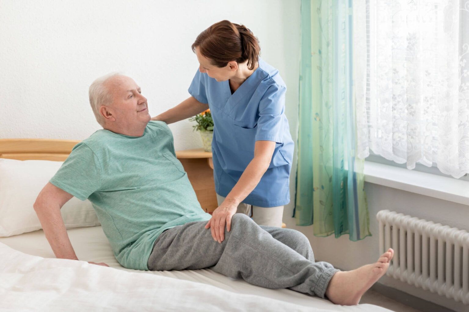 Caregiver helping a man sit up on a bed in a bright room