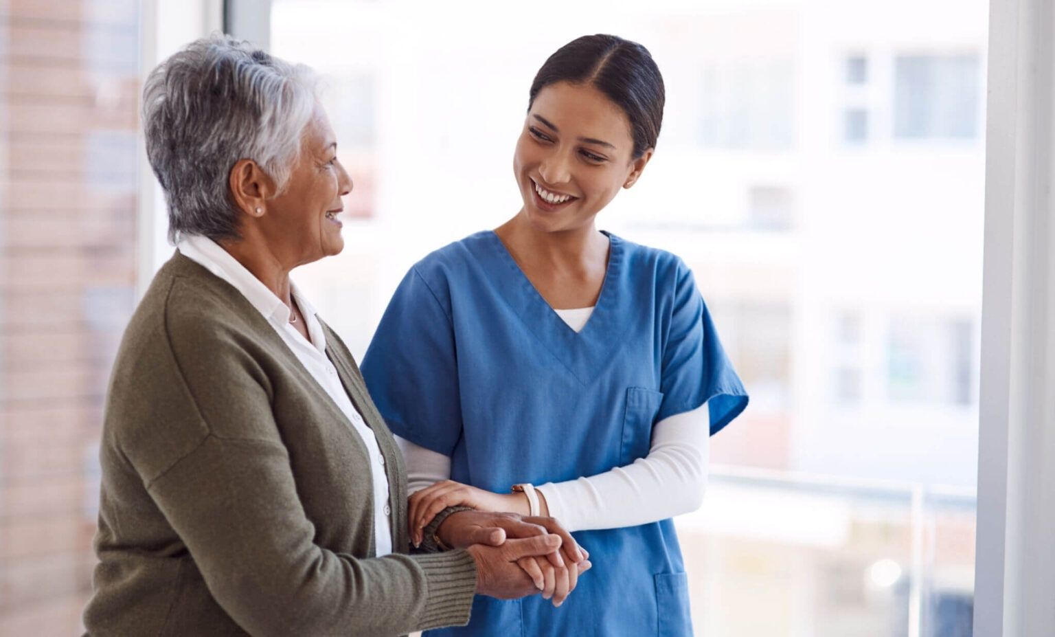 Nurse in blue scrubs gently holding hands with an elderly patient in a bright clinic setting