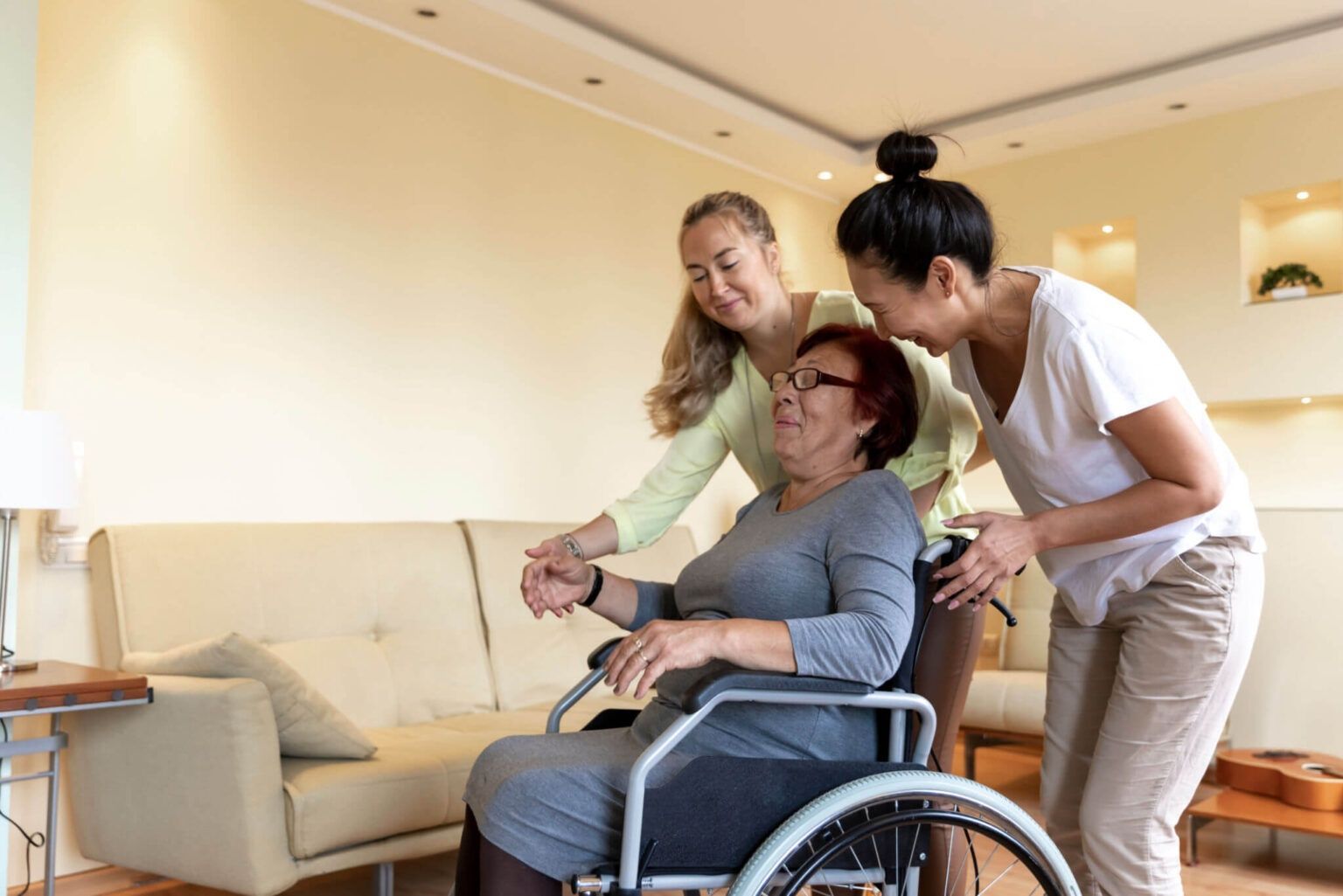 Two caregivers assist an older woman in a wheelchair in a bright living room.