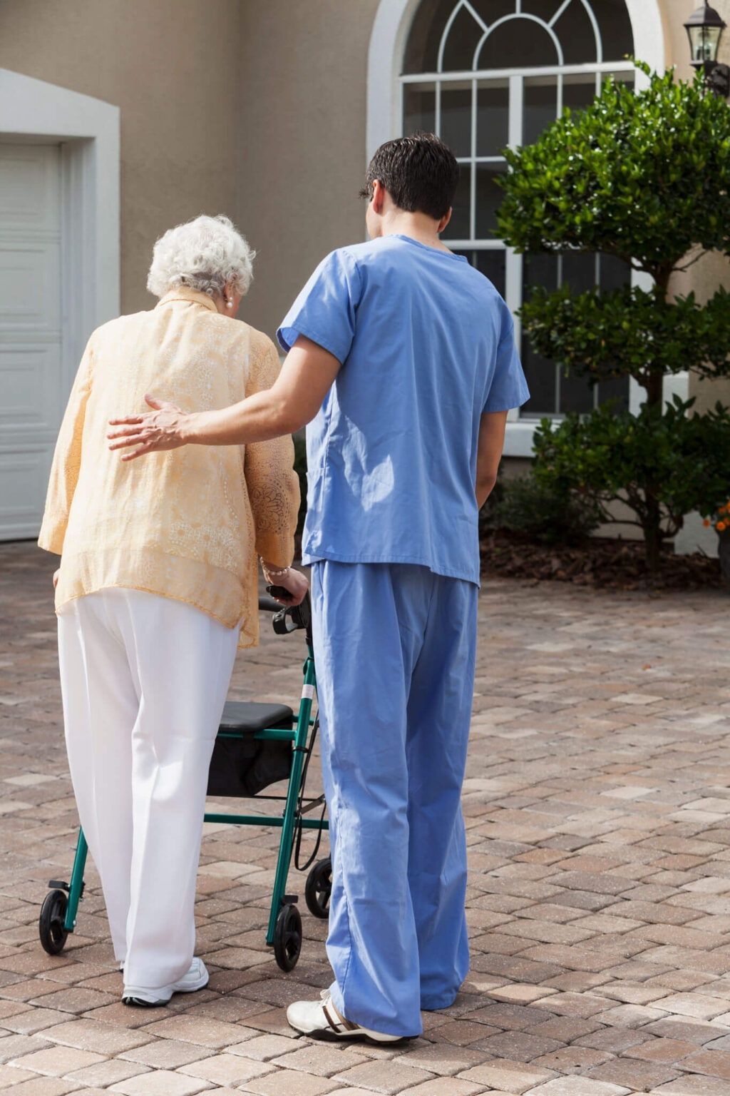 Nurse in blue scrubs guiding an elderly person with a walker along a house entry path