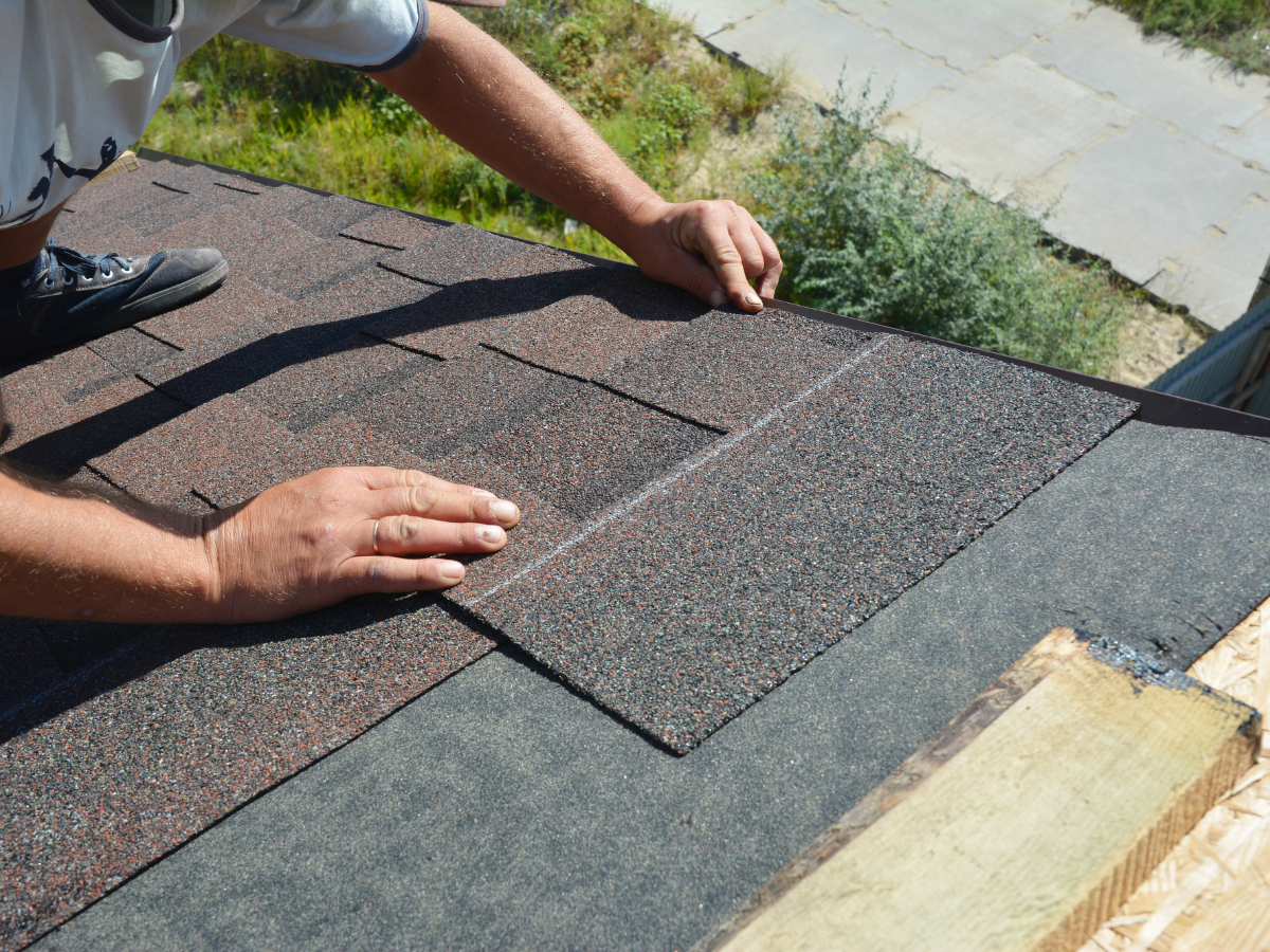 A person installing dark brown asphalt shingles on a roof over gray roofing felt.