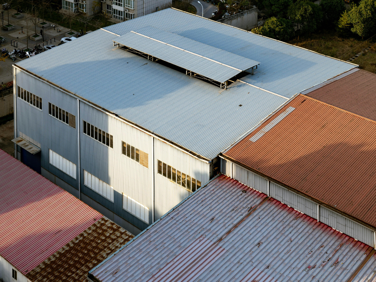 An aerial view of industrial buildings with corrugated metal and terracotta tile roofs under sunlight.