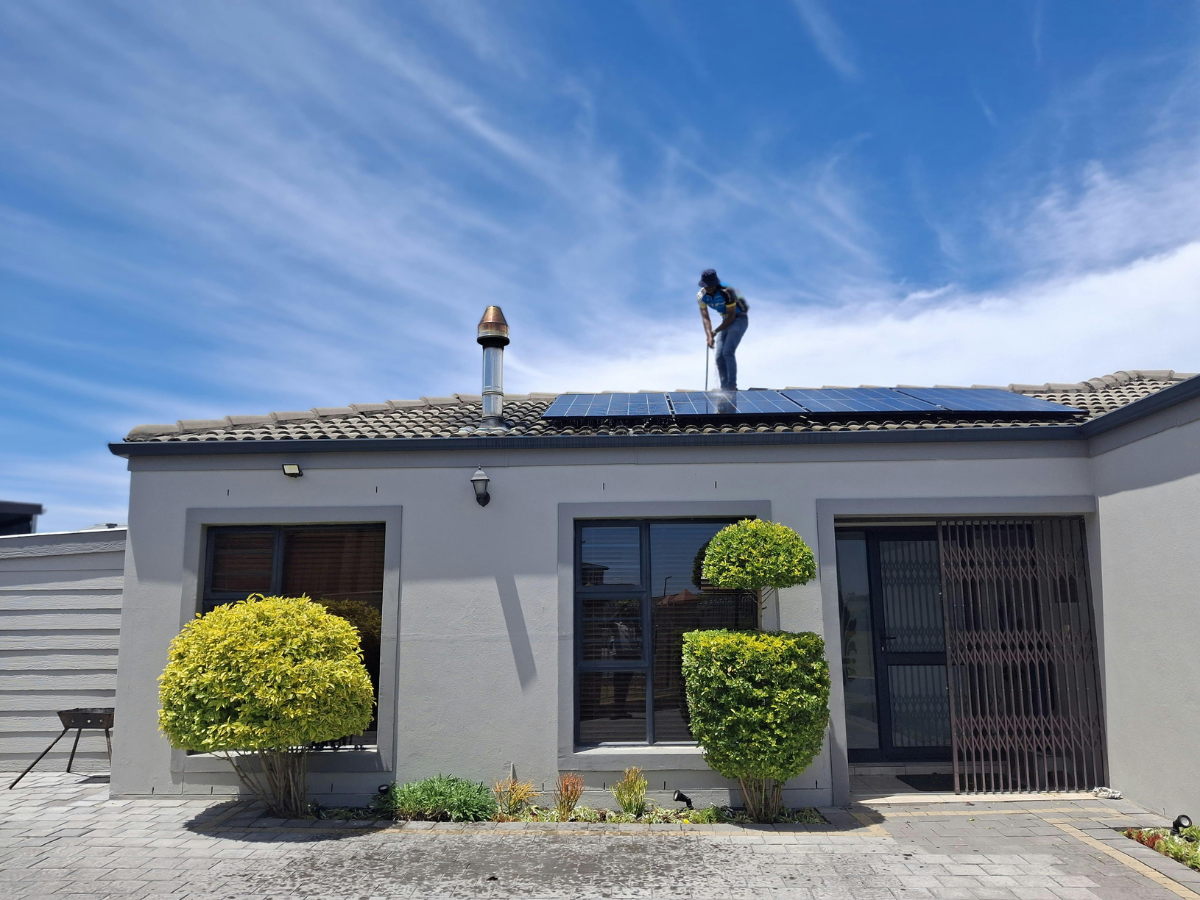A person stands on a house roof next to a solar panel array against a bright blue sky with wispy clouds.