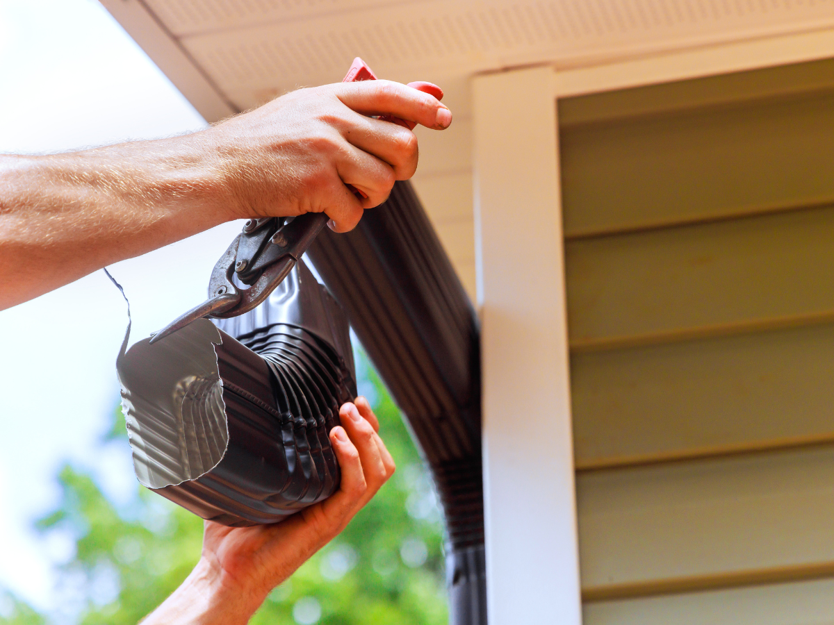 Close-up of hands connecting a brown downspout elbow piece to a gutter system on the side of a house.