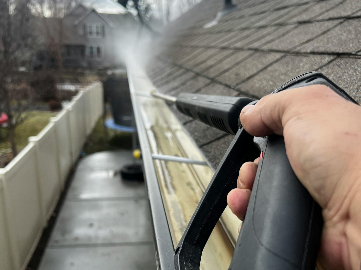 A first-person view of a person using a pressure washer wand to clean debris out of a residential rain gutter.