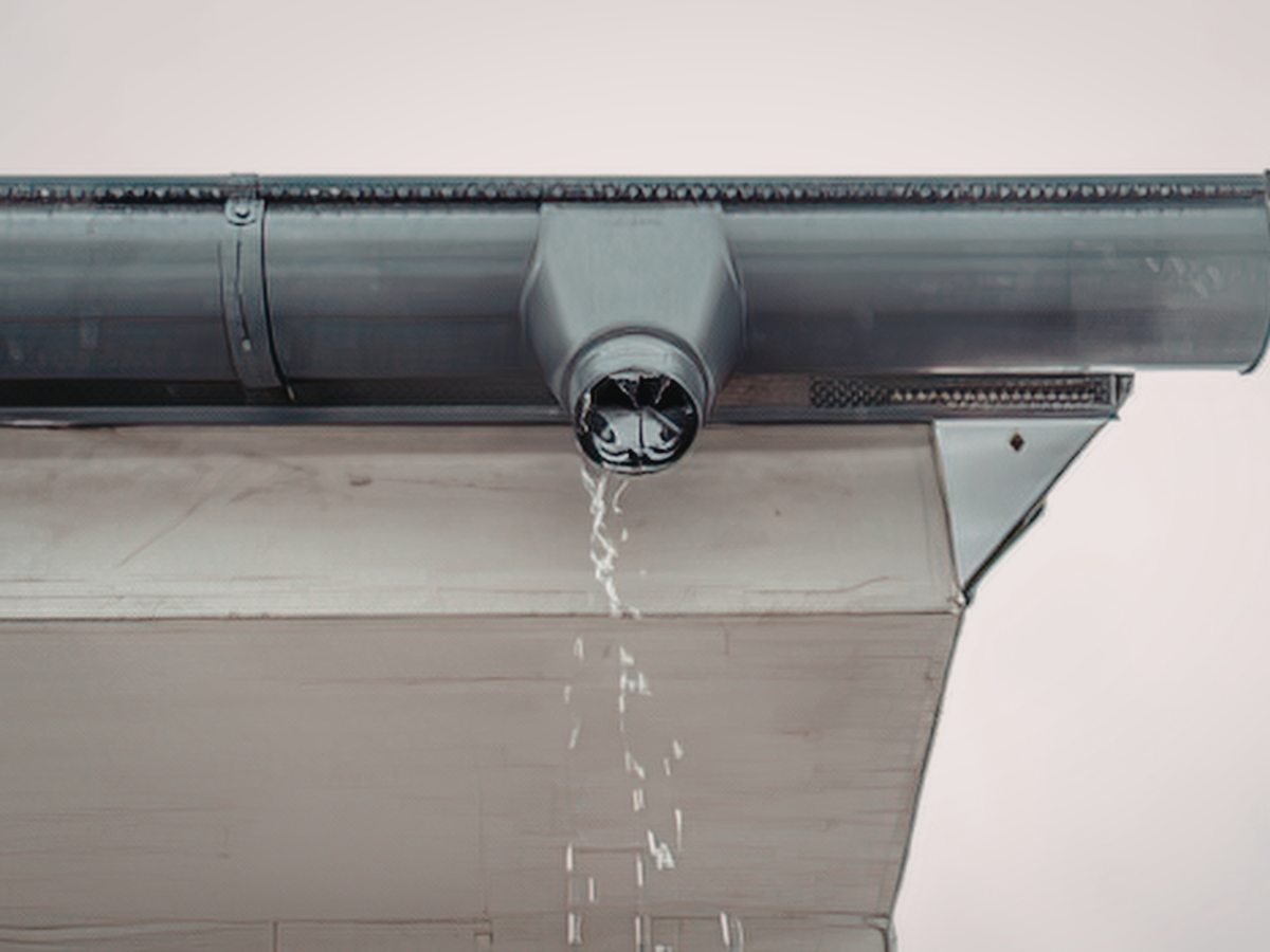 Water drips from the opening of a dark gray gutter attached to a building's fascia board.