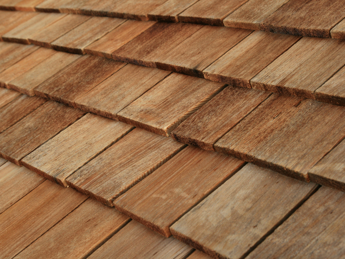 A close-up view of overlapping wooden roof shingles, showing natural brown wood grain and textures in a repeating pattern.