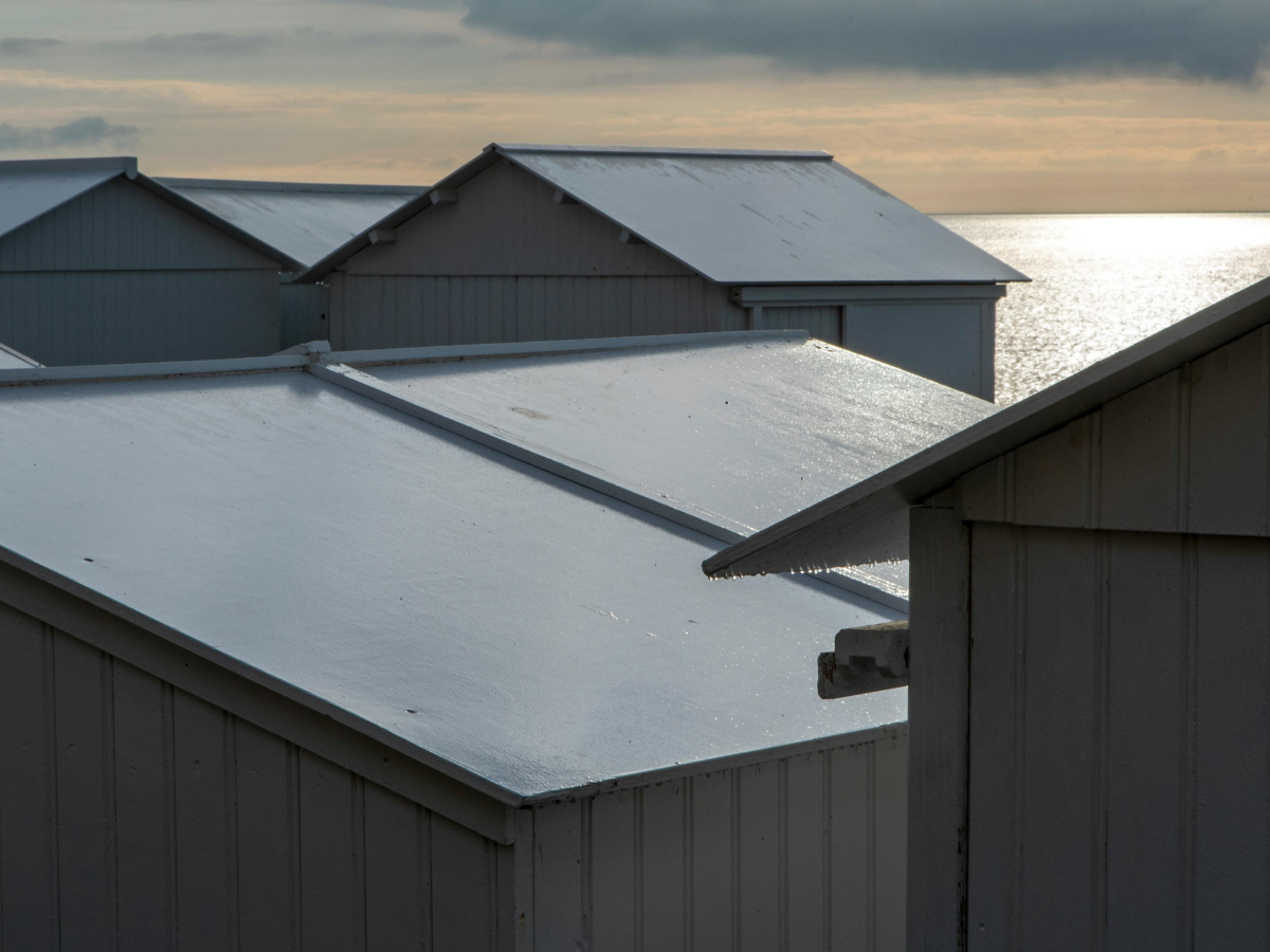 Light gray beach huts stand together under a cloudy sky, with the ocean reflecting sunlight in the background.