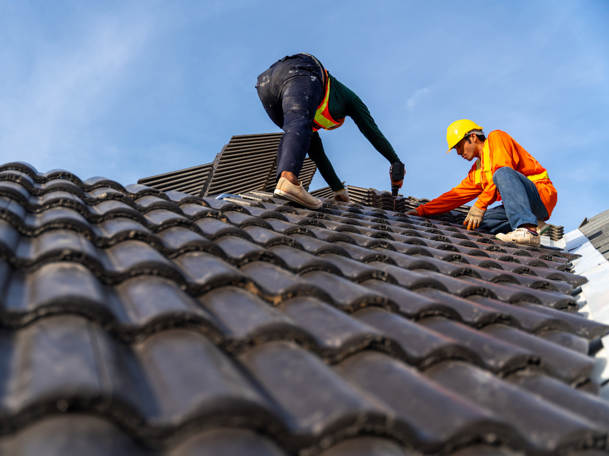 Two workers in safety gear and hard hats install dark, curved roof tiles against a clear blue sky.