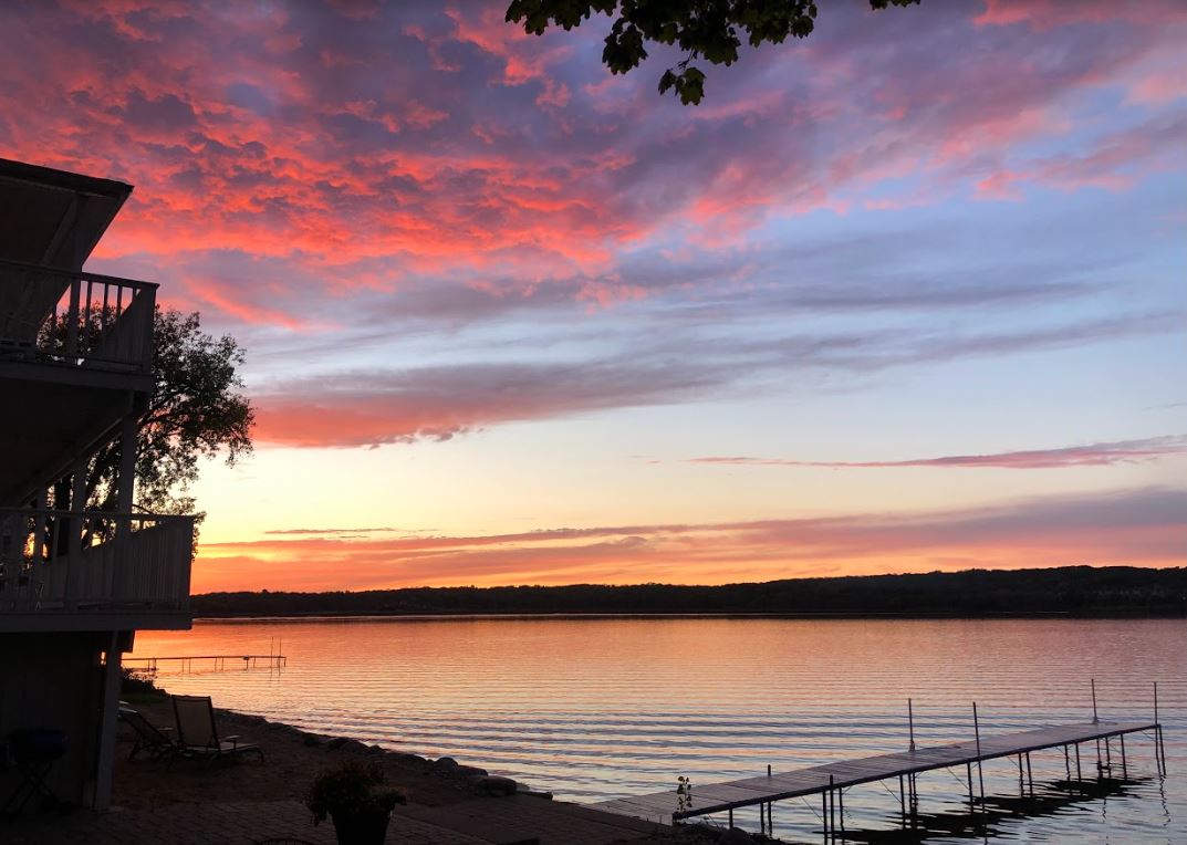 A sunset over a lake with a dock in the foreground and a house in the background.